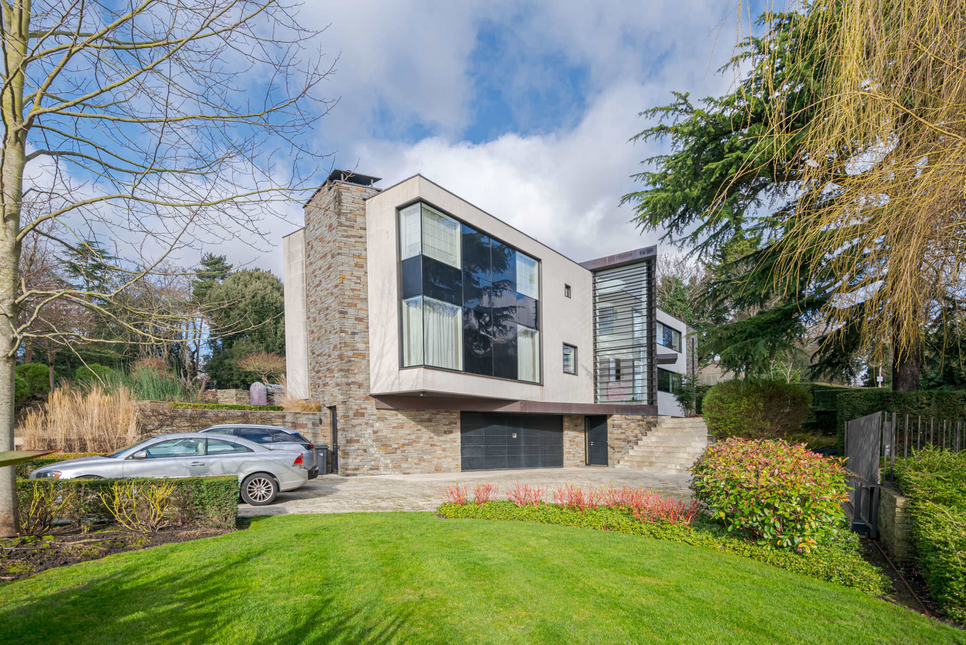 A modern, two-story house featuring large windows and a stone facade is set amidst landscaped greenery and a driveway.