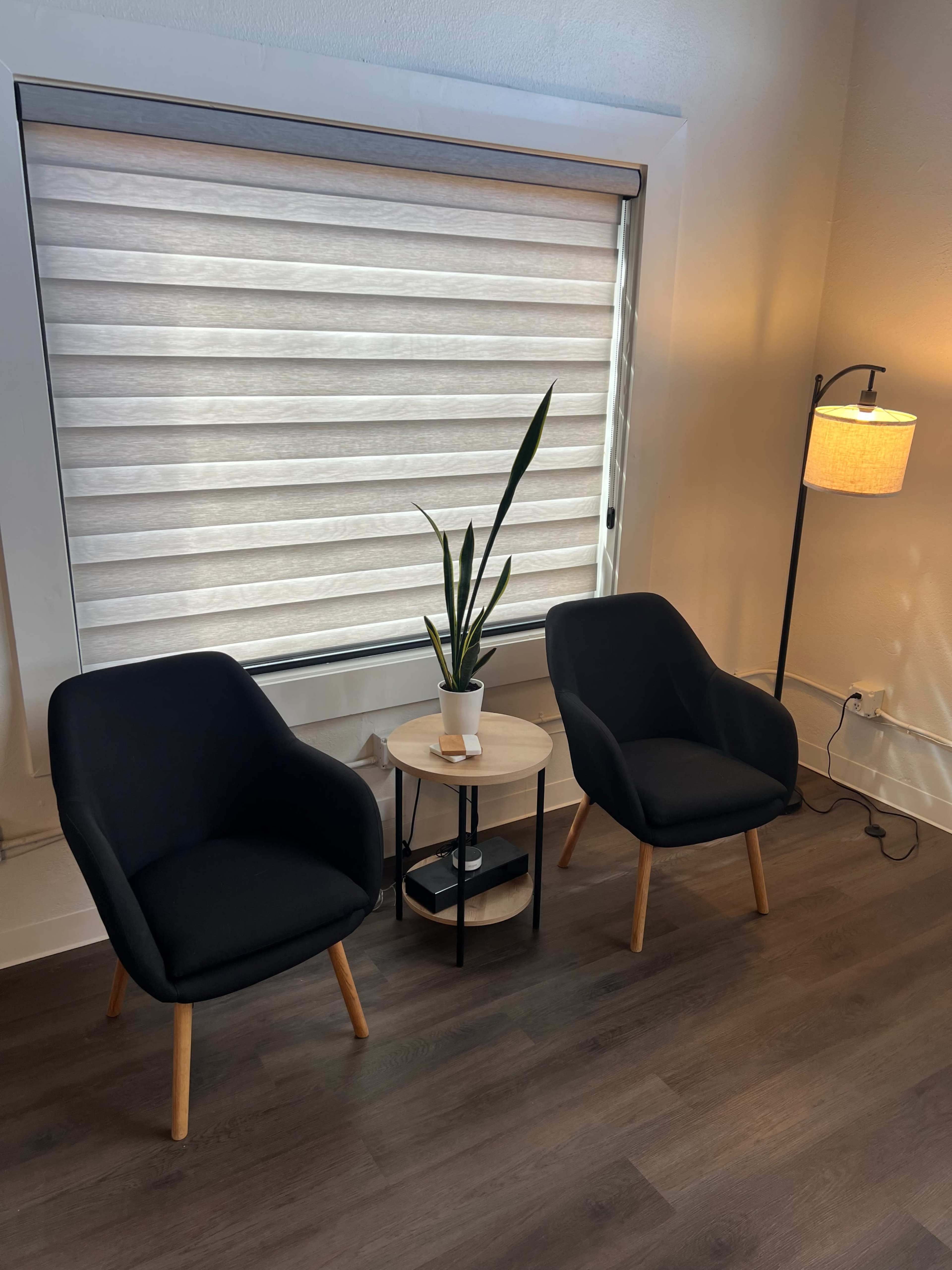 The image shows a cozy seating area with two black chairs, a small circular table, a potted plant, and a floor lamp beside a window with light-filtering shades.