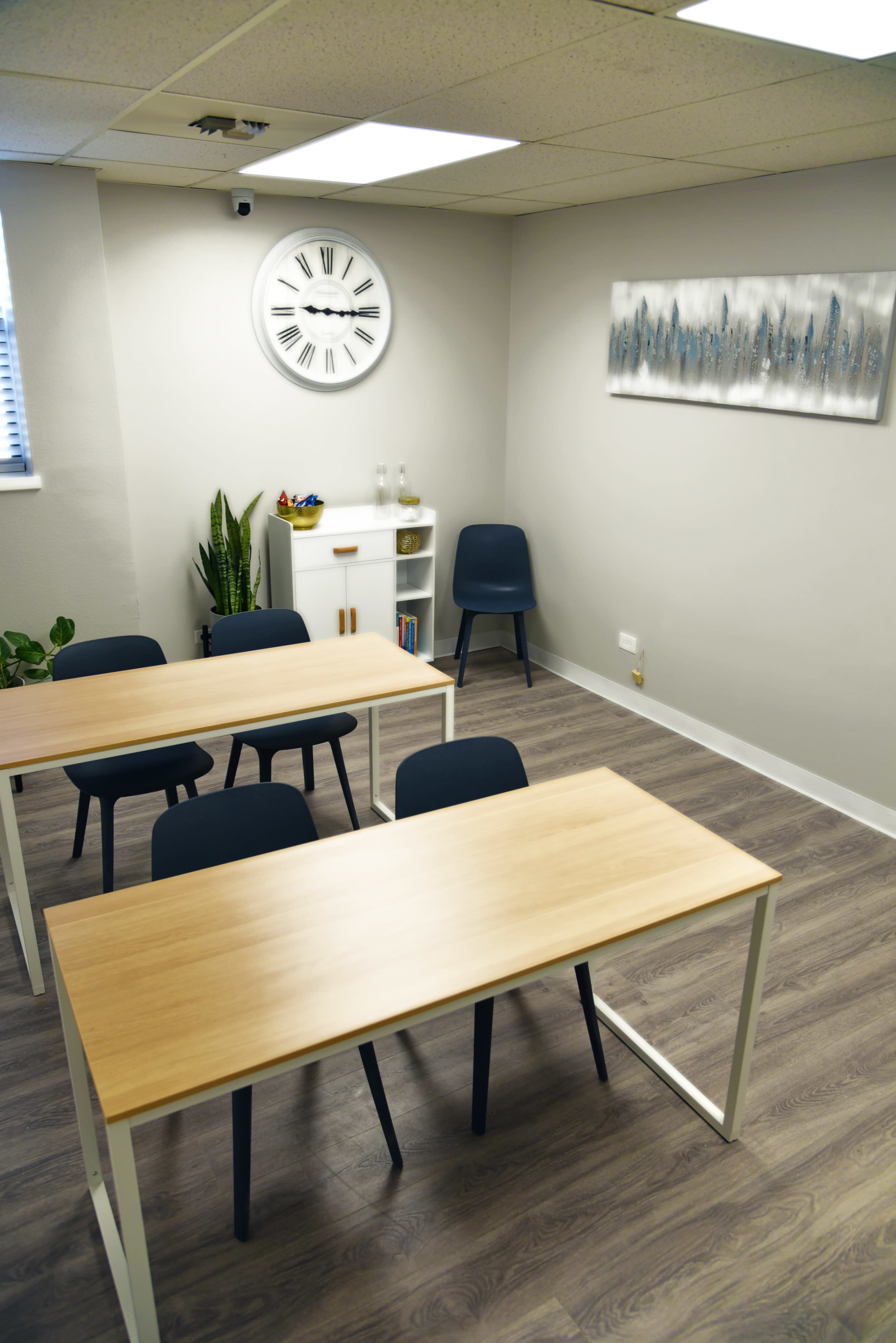 A small meeting room with four chairs and two tables, a clock on the wall, and a small cabinet beside a plant.