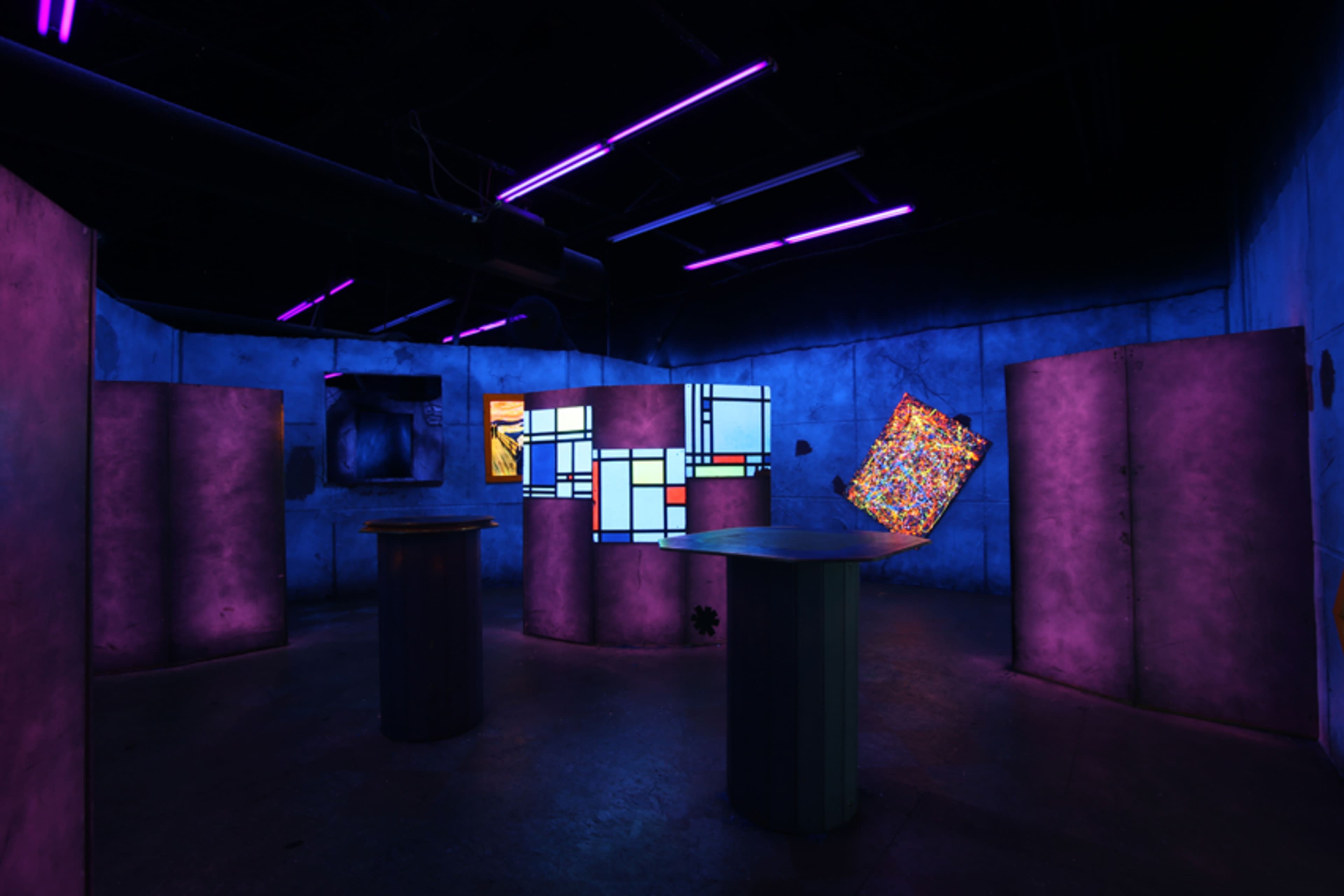 The image shows an art installation in a dimly lit room with walls illuminated by purple and blue lights, featuring various abstract displays and columns.
