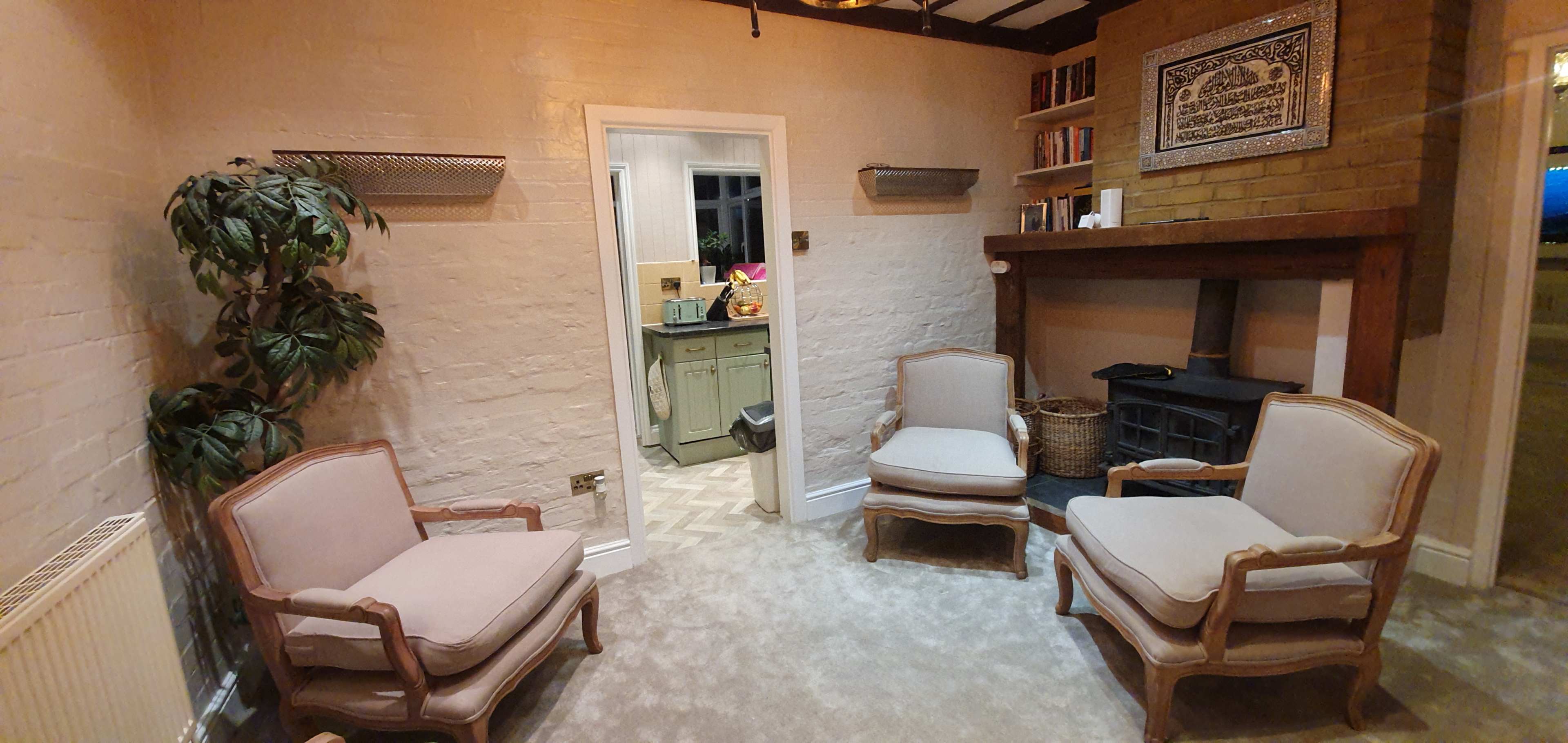 A cozy living room with three beige armchairs arranged around a central area, a fireplace against a brick wall, and a doorway leading to another room.