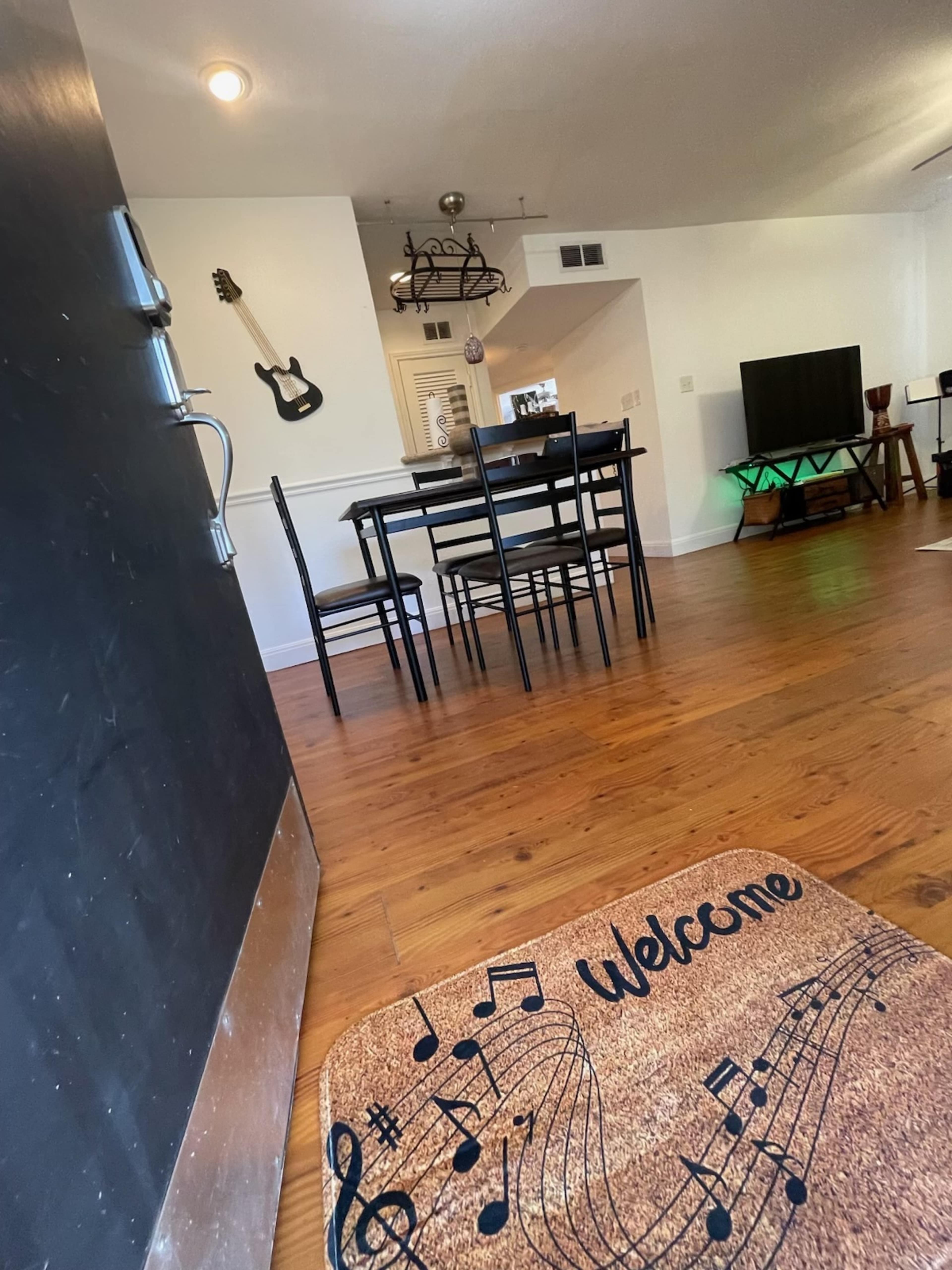 A welcome mat with musical notes lies at the entrance of a brightly lit room featuring a dining table with chairs, a television, and a guitar hanging on the wall.