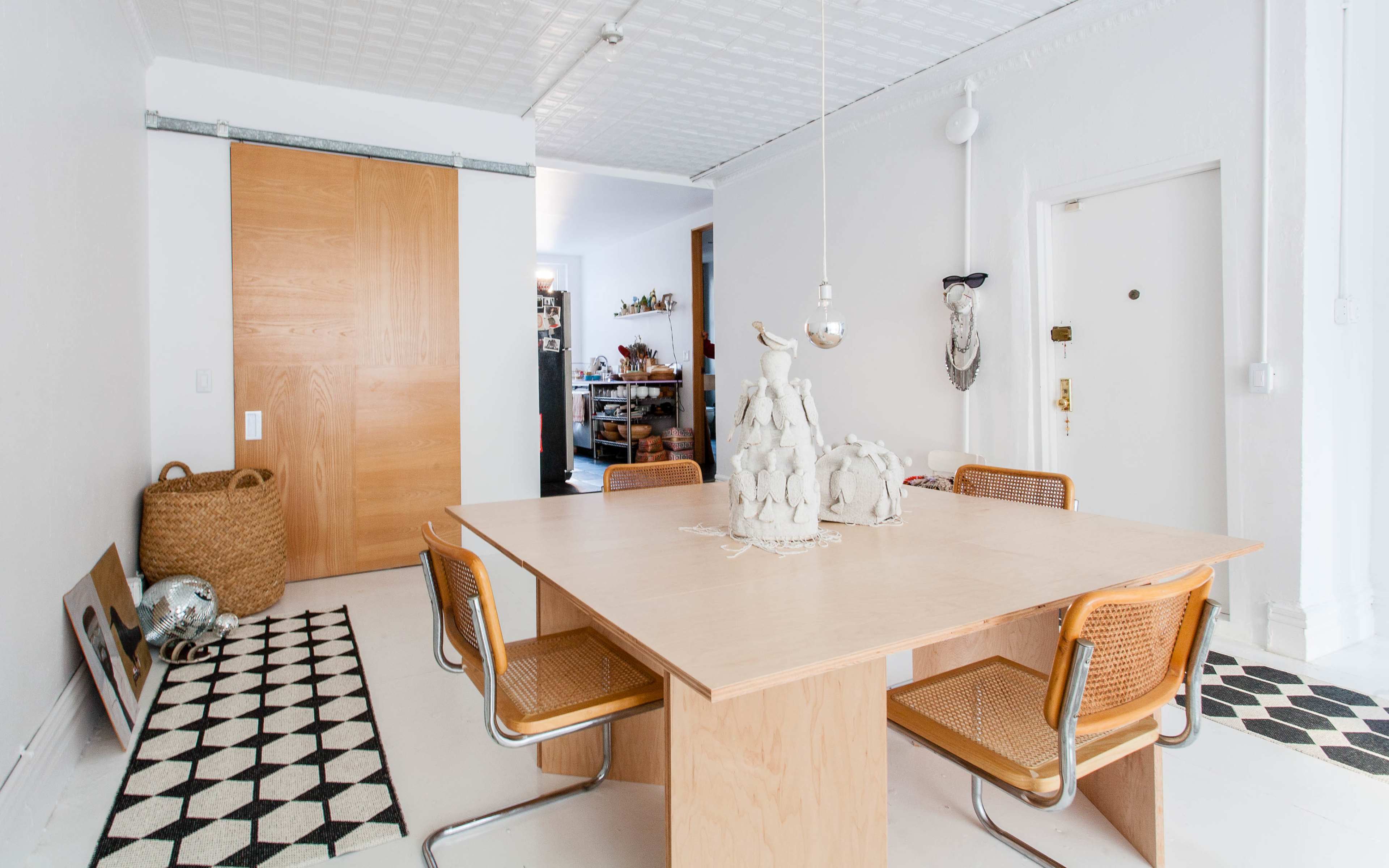 A minimalistic dining area with a wooden table surrounded by woven chairs, a patterned rug, and a sliding wooden door in the background.