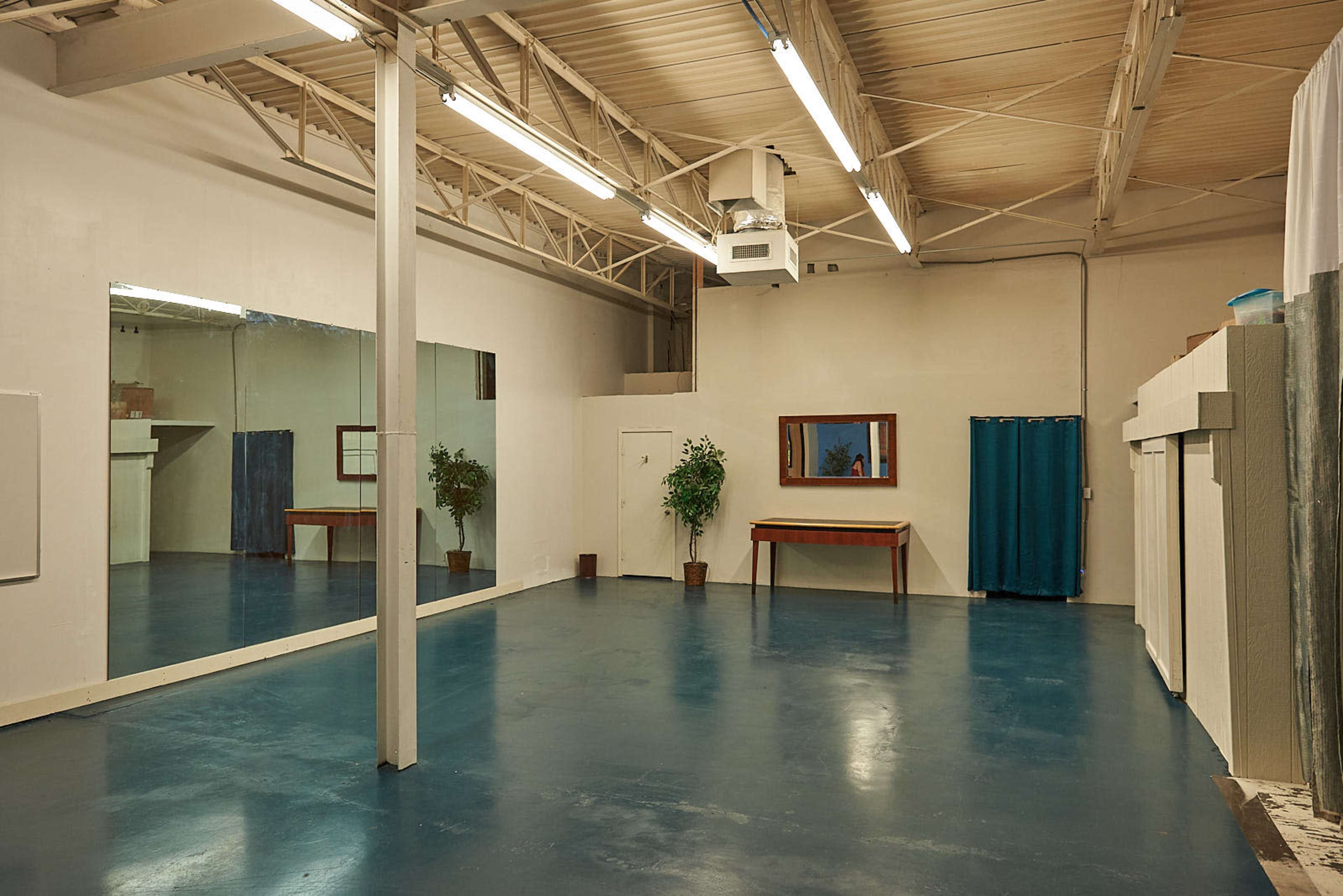 The room features a large mirrored wall, a wooden table against one wall, and a door leading to another space, all set on a glossy blue floor.