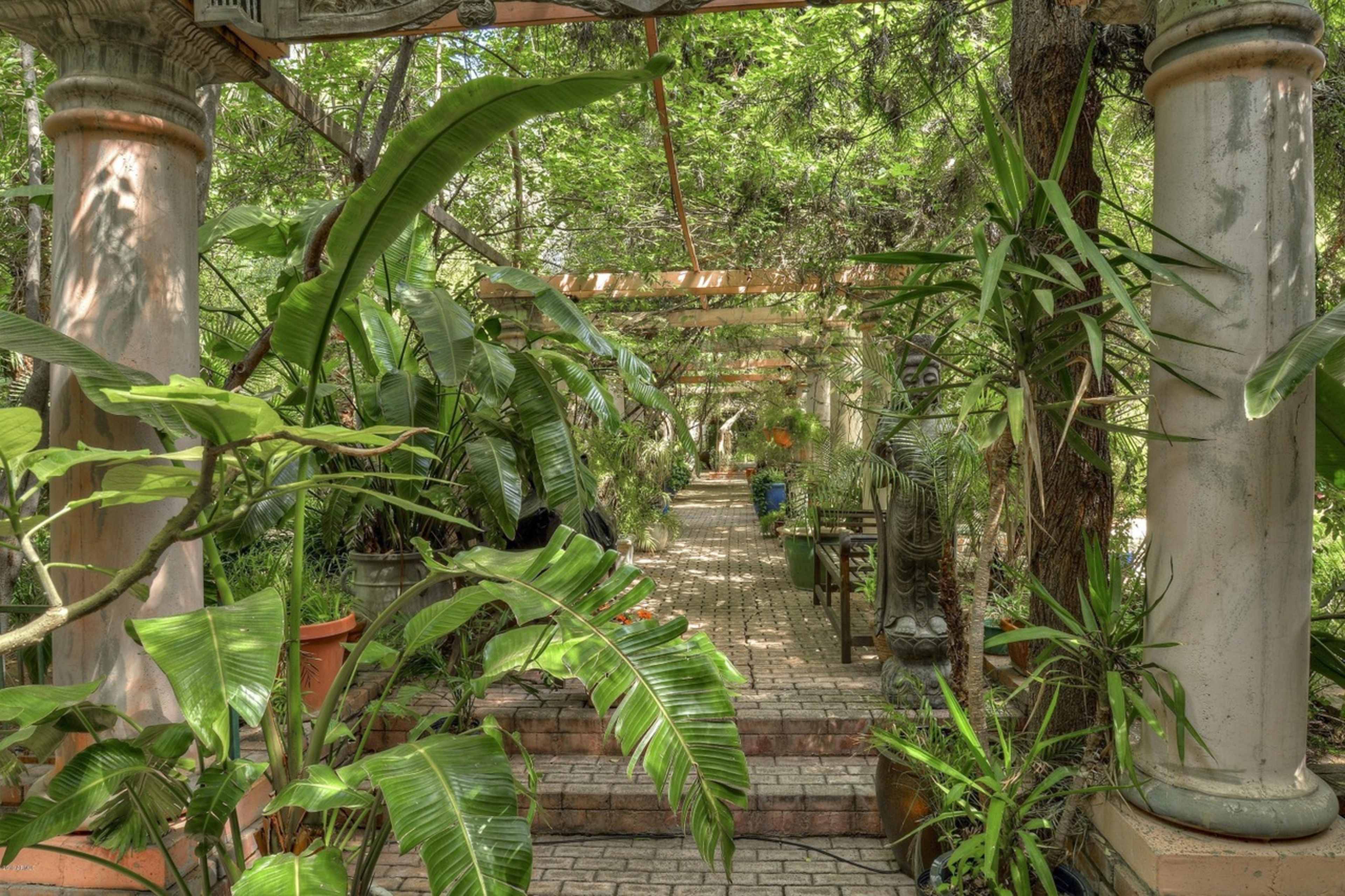 The image shows a landscaped garden pathway lined with columns and lush greenery, leading deeper into the space.