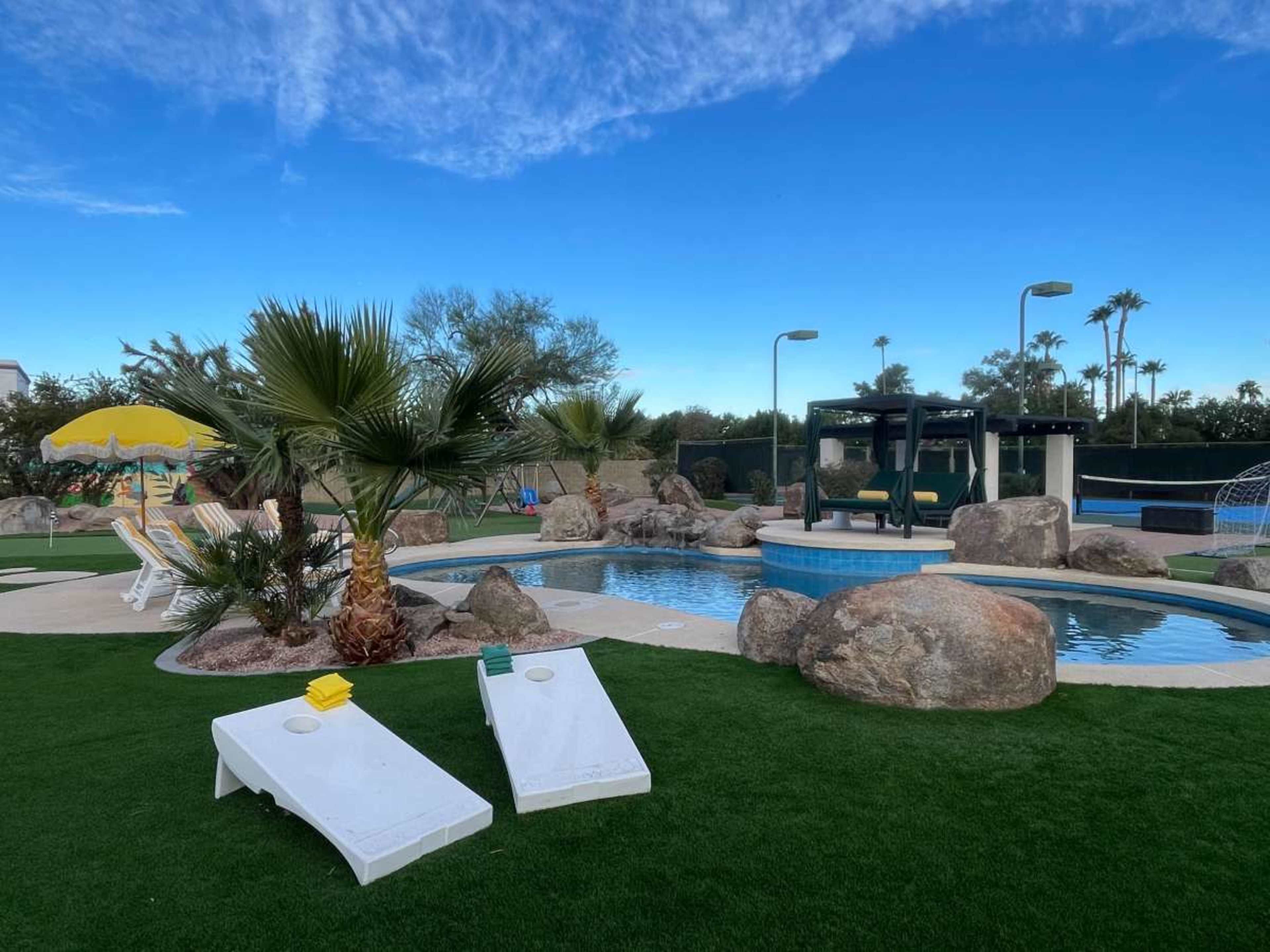 The image shows a landscaped pool area featuring two white lounge chairs, palm trees, large rocks, and a bright blue sky with a few clouds.
