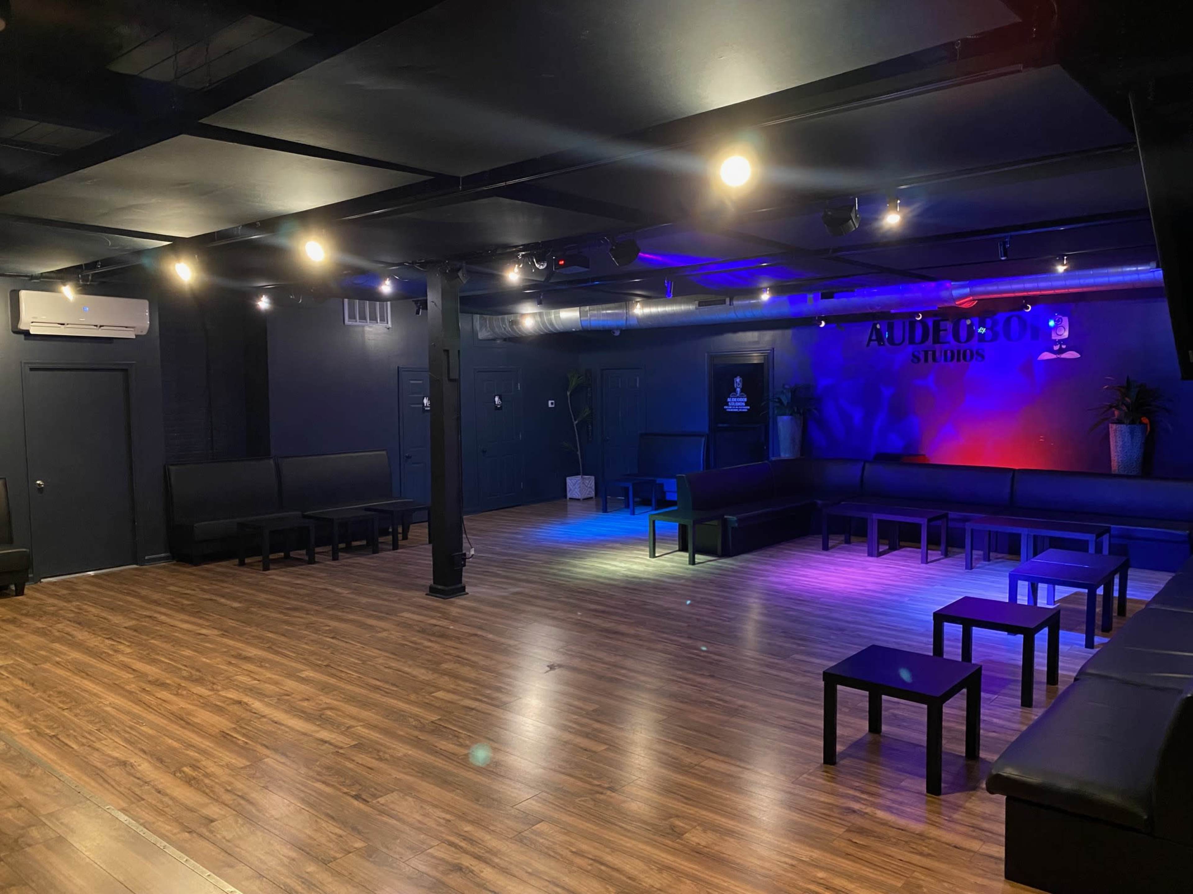 A spacious studio with dark walls, wooden flooring, and black seating arranged around small tables, illuminated by colorful lights.