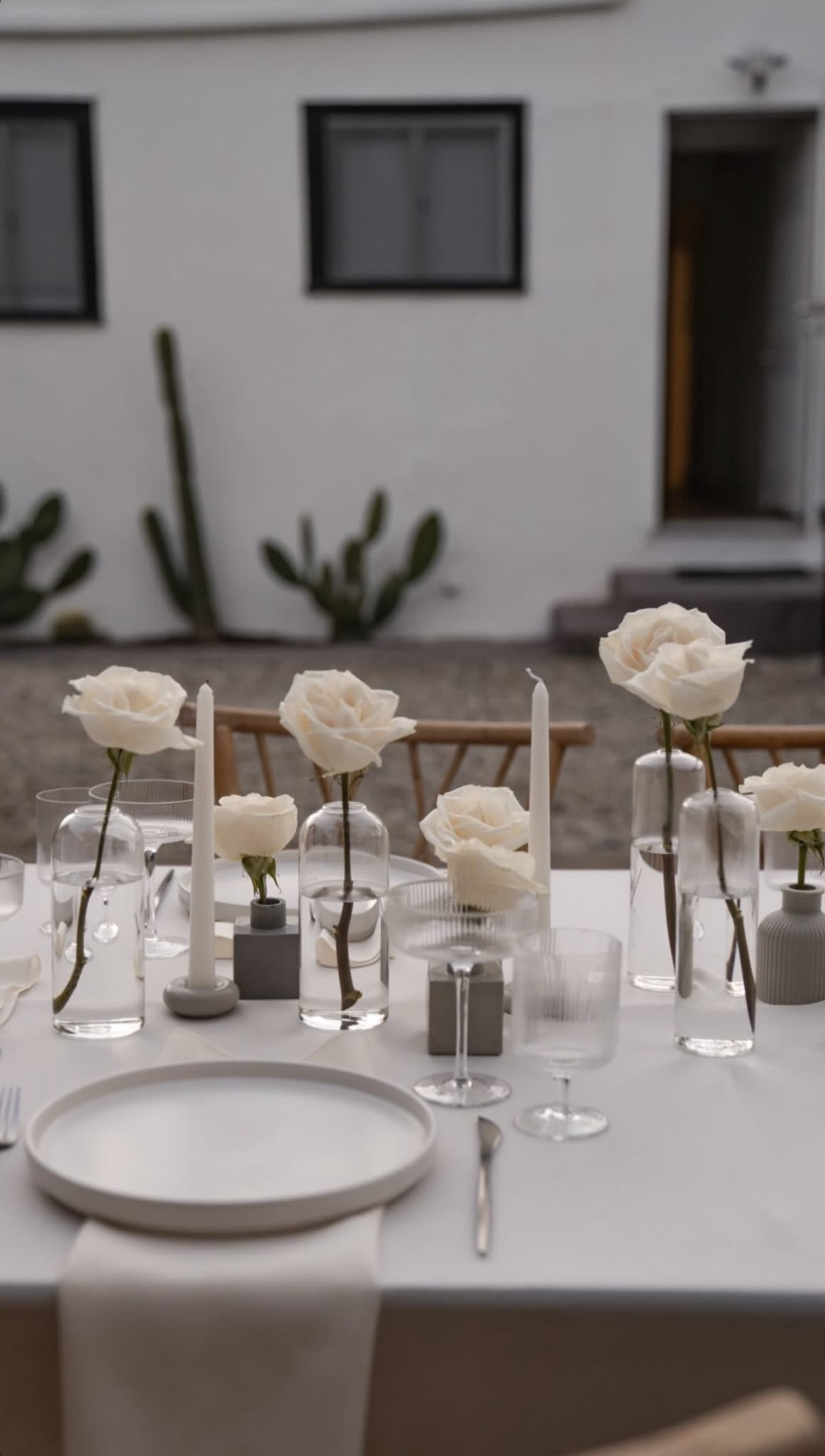 A styled outdoor dining table is set with white roses in glass vases, candles, and elegant tableware, against a backdrop of a white building and cacti.