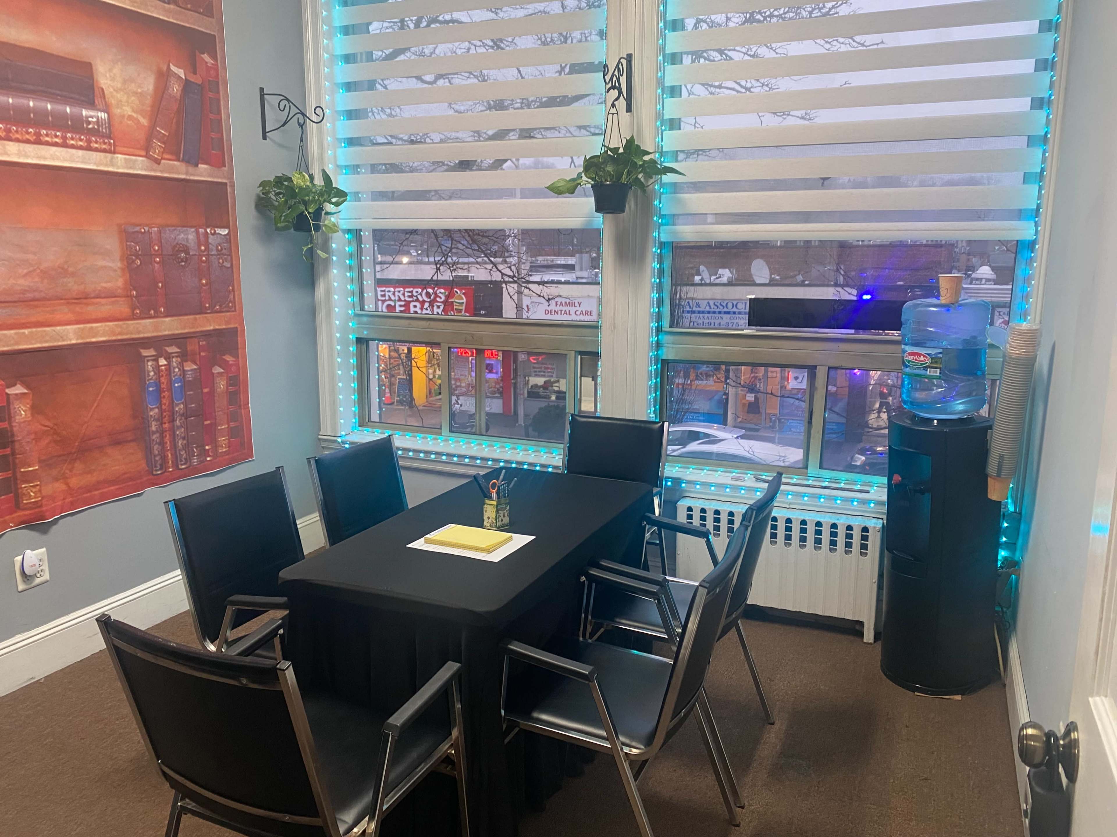 A small meeting room features a black table surrounded by six chairs, with a water cooler and large windows displaying a street view.