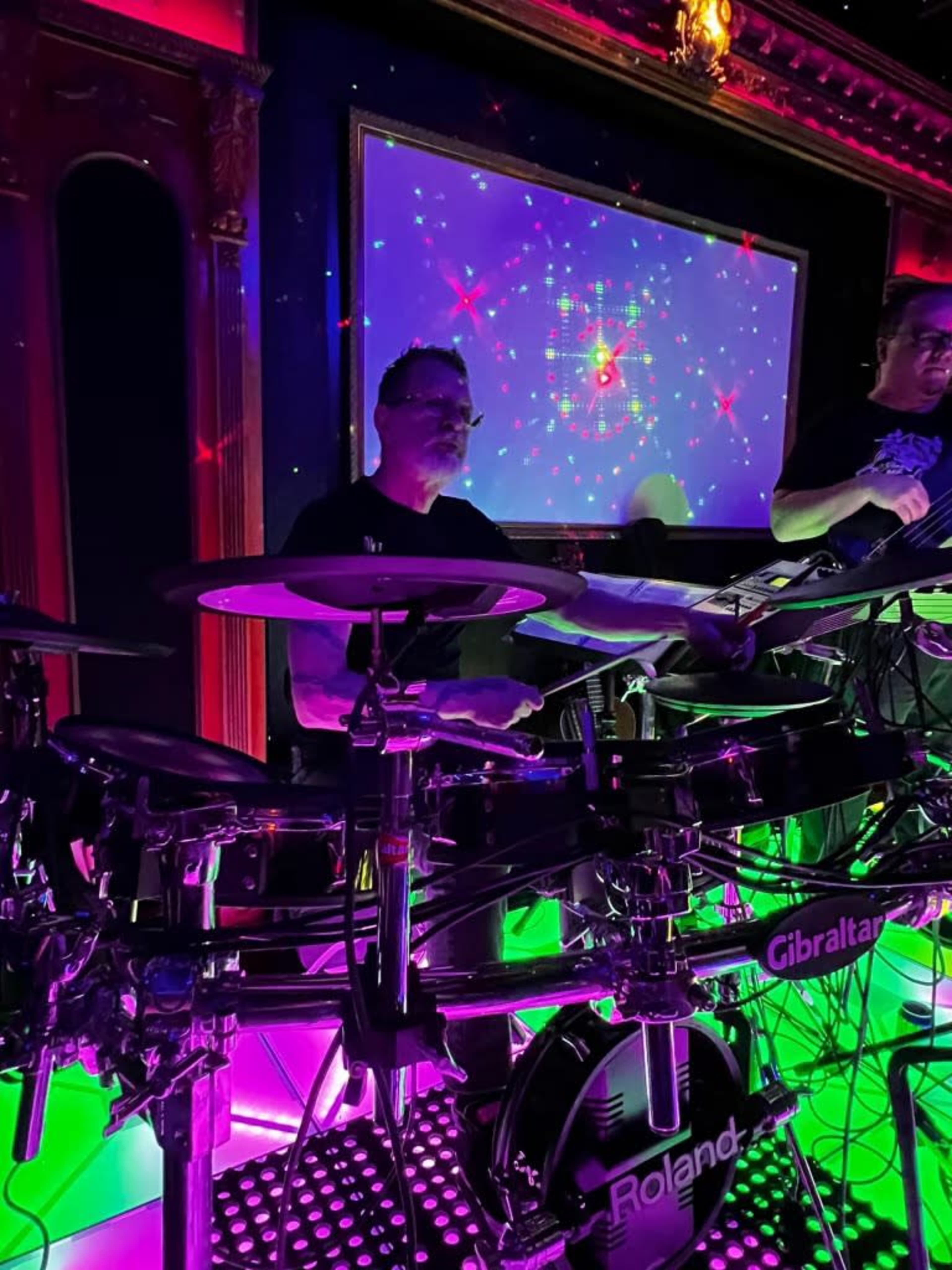 A drummer plays an electronic drum set on a brightly lit stage with a colorful backdrop.