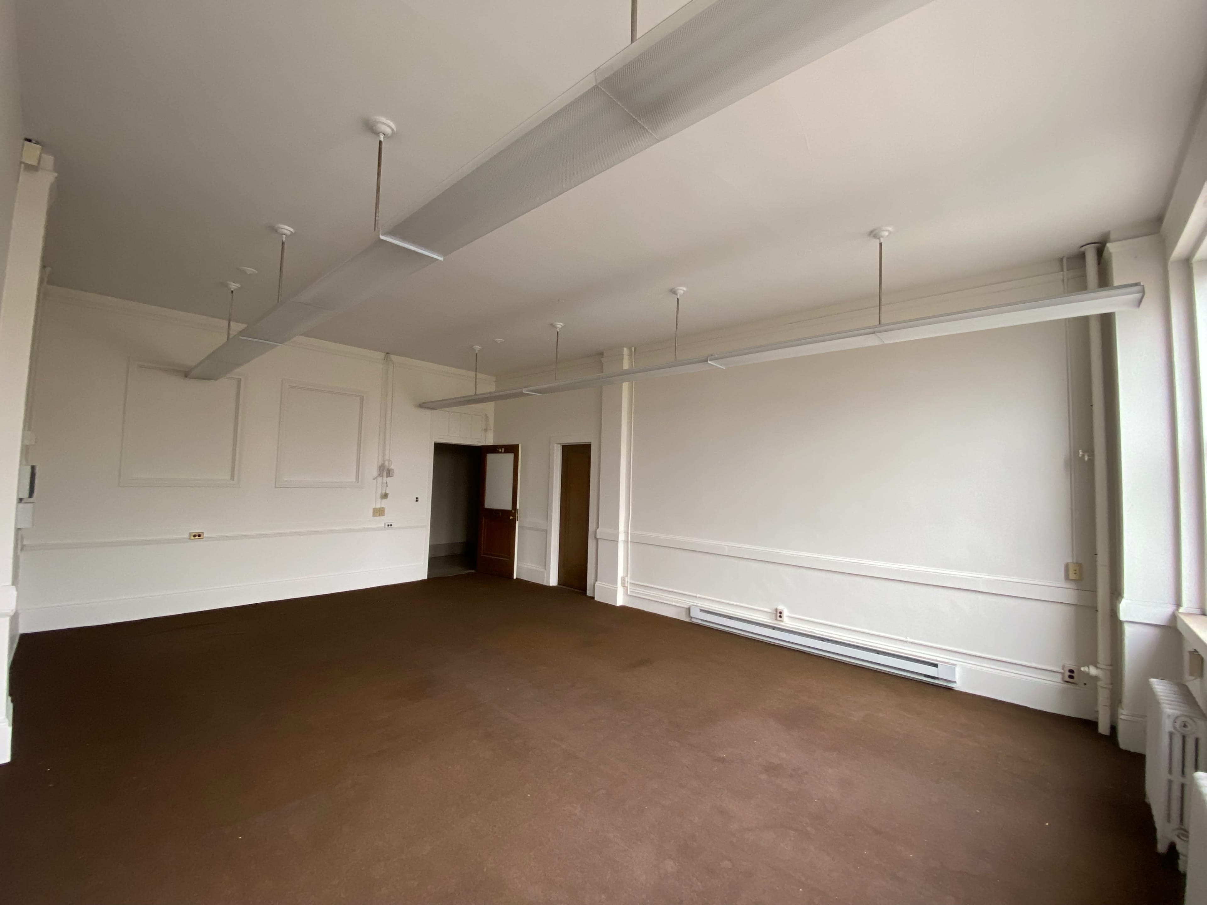 The image shows a spacious, empty room with beige walls, a brown floor, and a ceiling fitted with lights.