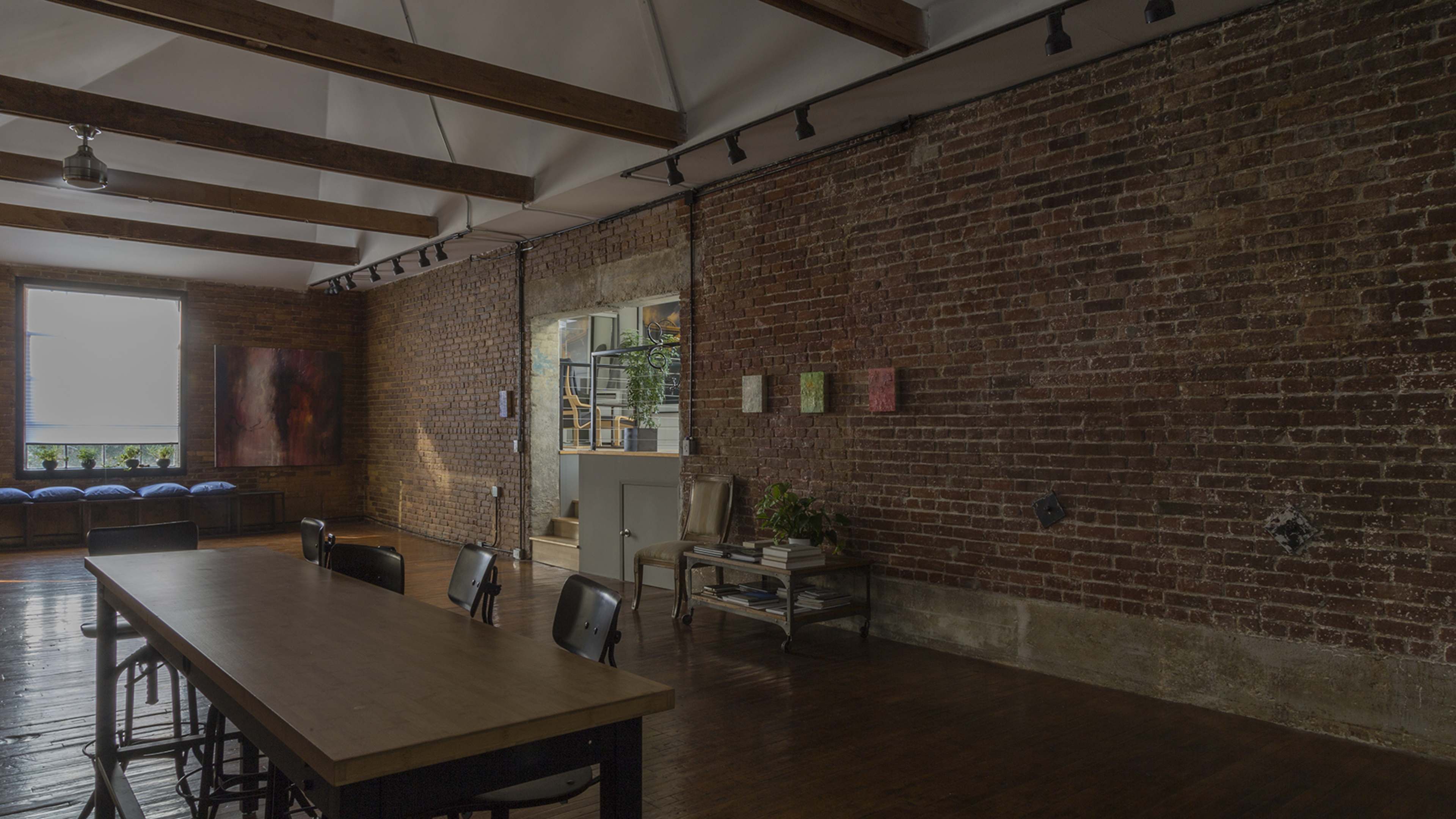 A spacious room with exposed brick walls, a long wooden table, and natural light streaming in from a window.
