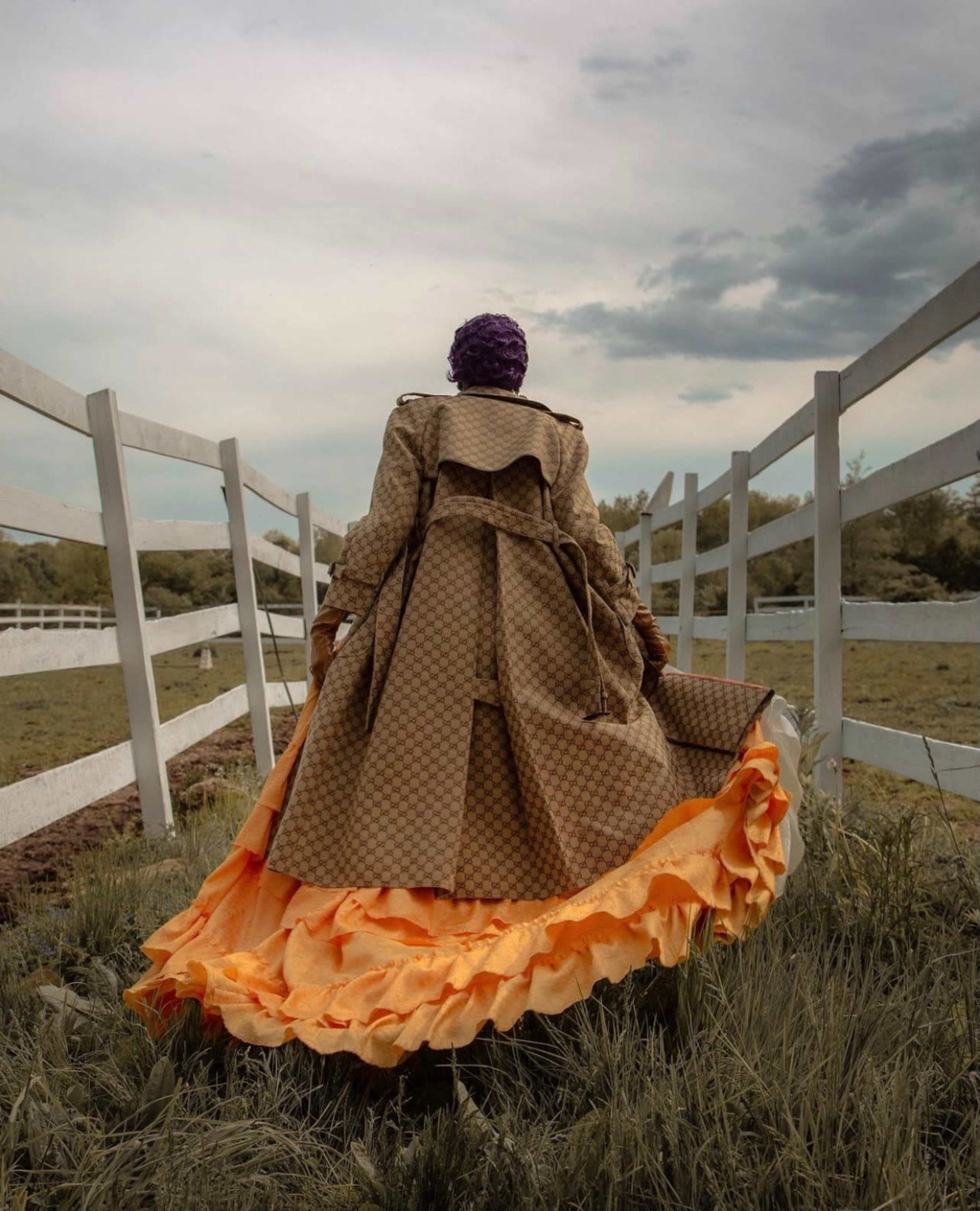 A person wearing a long coat and a brightly colored, ruffled dress stands in a grassy area surrounded by a white wooden fence under a cloudy sky.