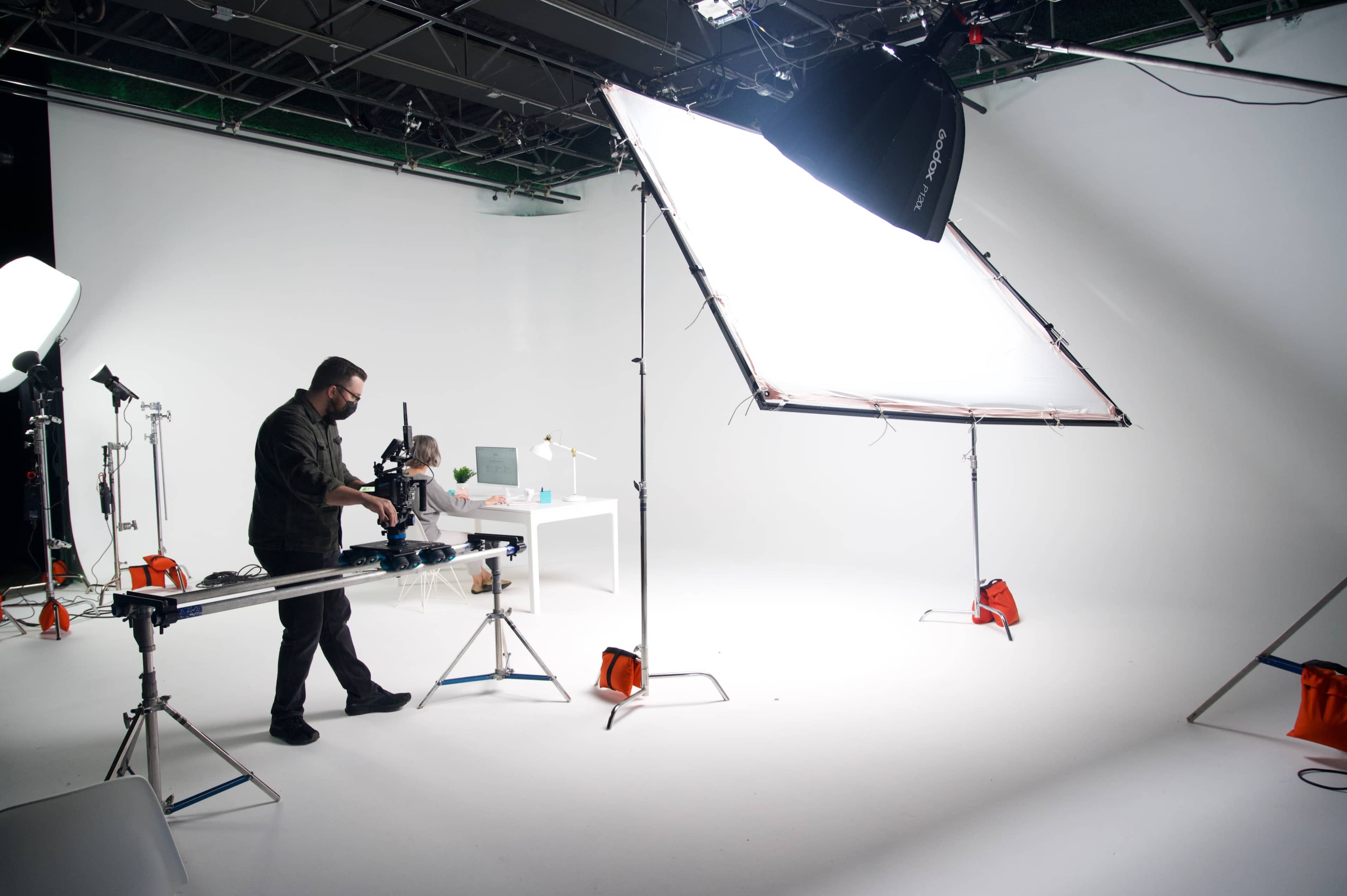 A photographer sets up equipment in a well-lit studio with a large light panel and a backdrop for a photoshoot.