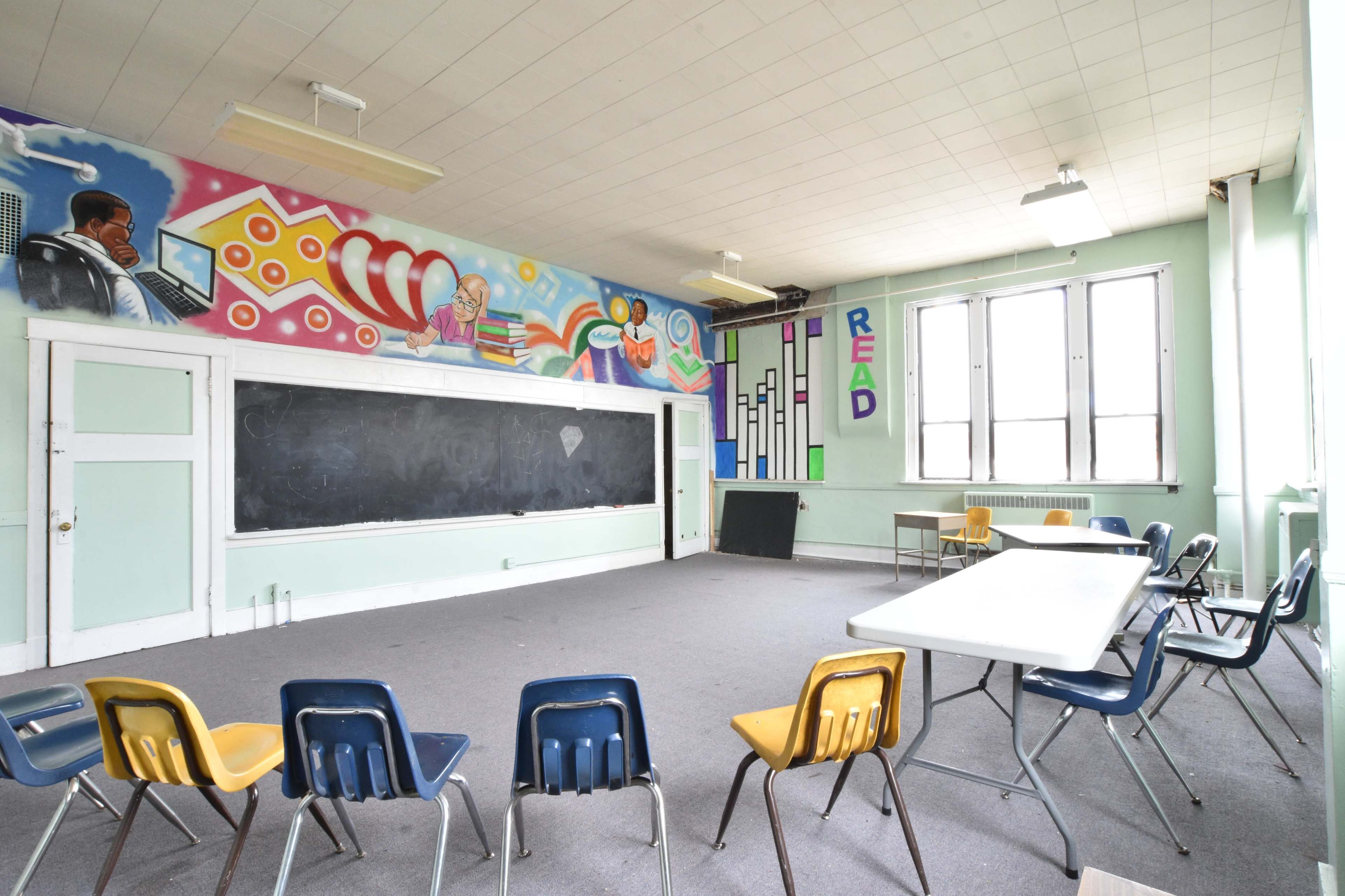 A classroom features a chalkboard at the front and a circular arrangement of chairs, with a vibrant mural on the walls.
