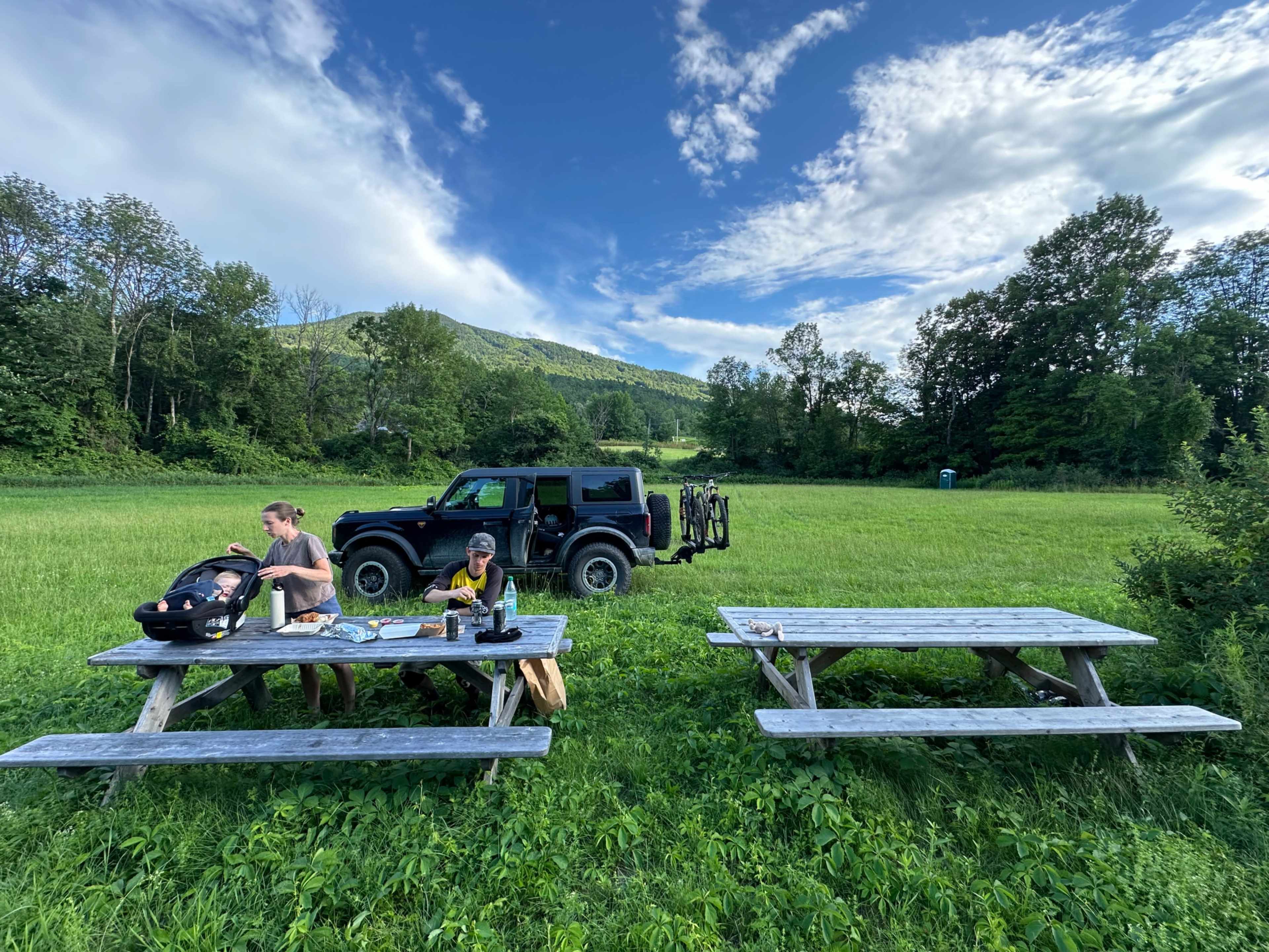 Two people sit at a picnic table with a baby carrier beside them, while a black Jeep is parked nearby in a grassy field surrounded by trees and hills.