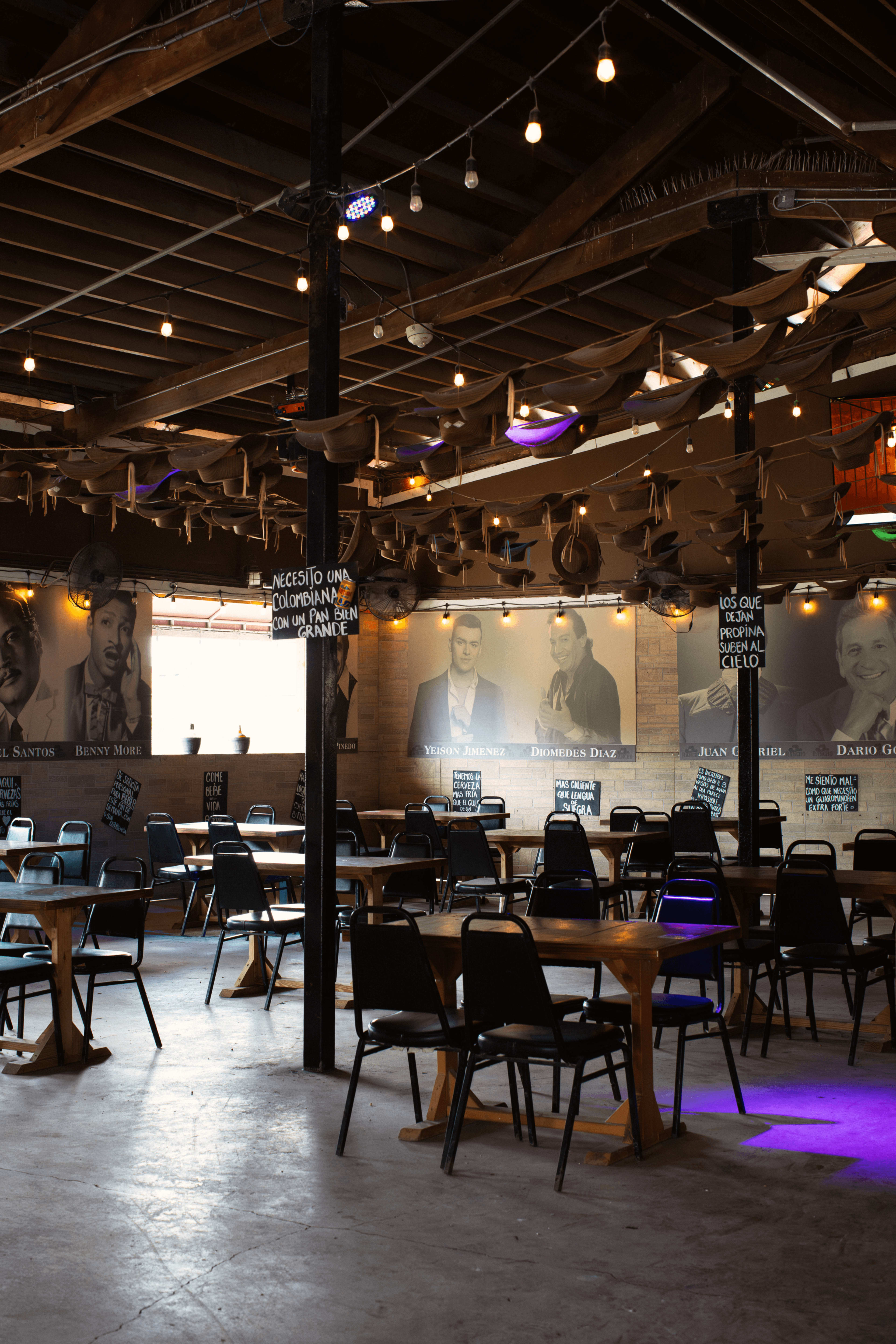 The image shows an indoor dining space with wooden tables, black chairs, and illuminated murals of people on the walls, under a ceiling decorated with light fixtures and draped materials.