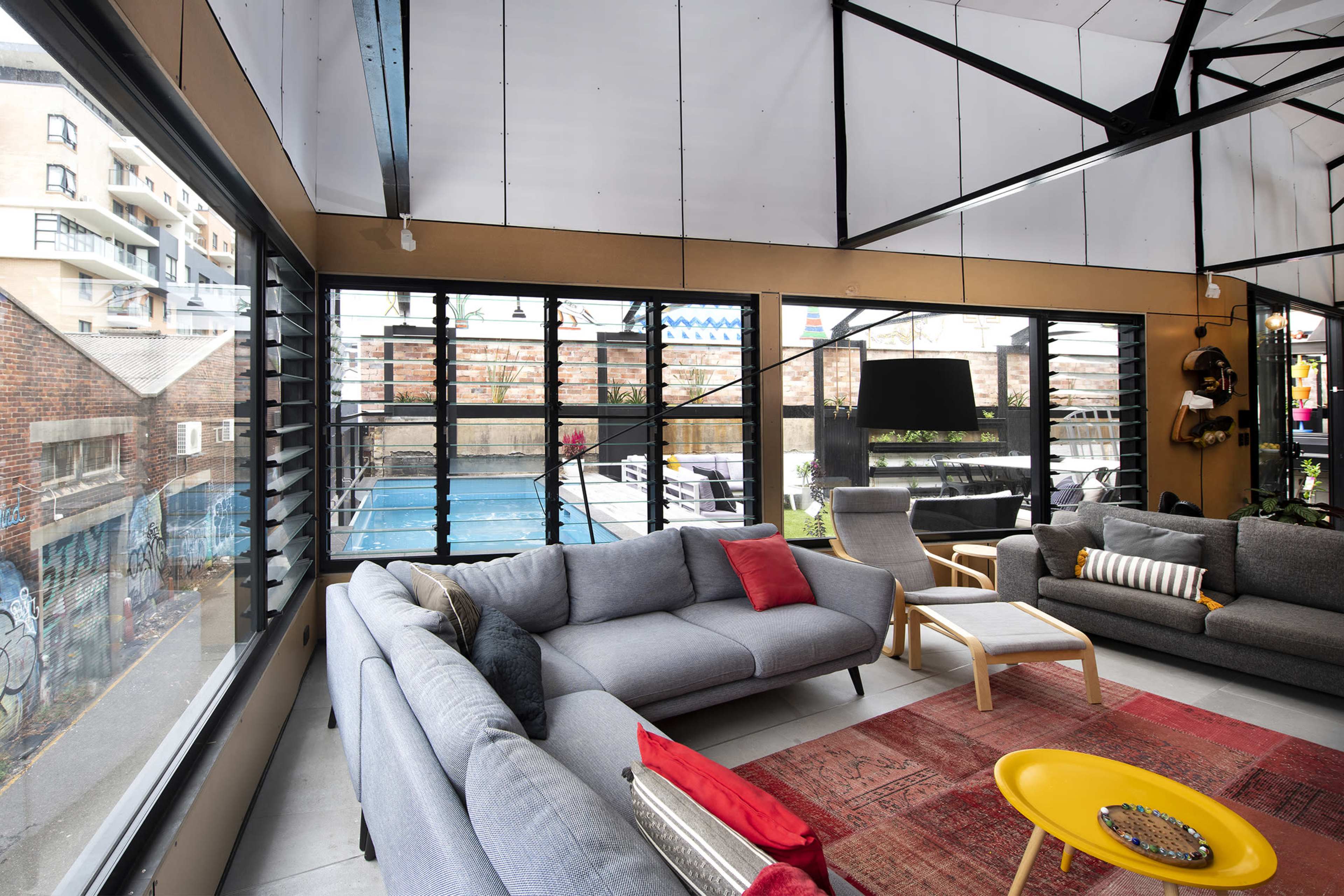 A modern living area with large windows that overlook a pool and outdoor seating, featuring gray sofas, a red rug, and a yellow coffee table.