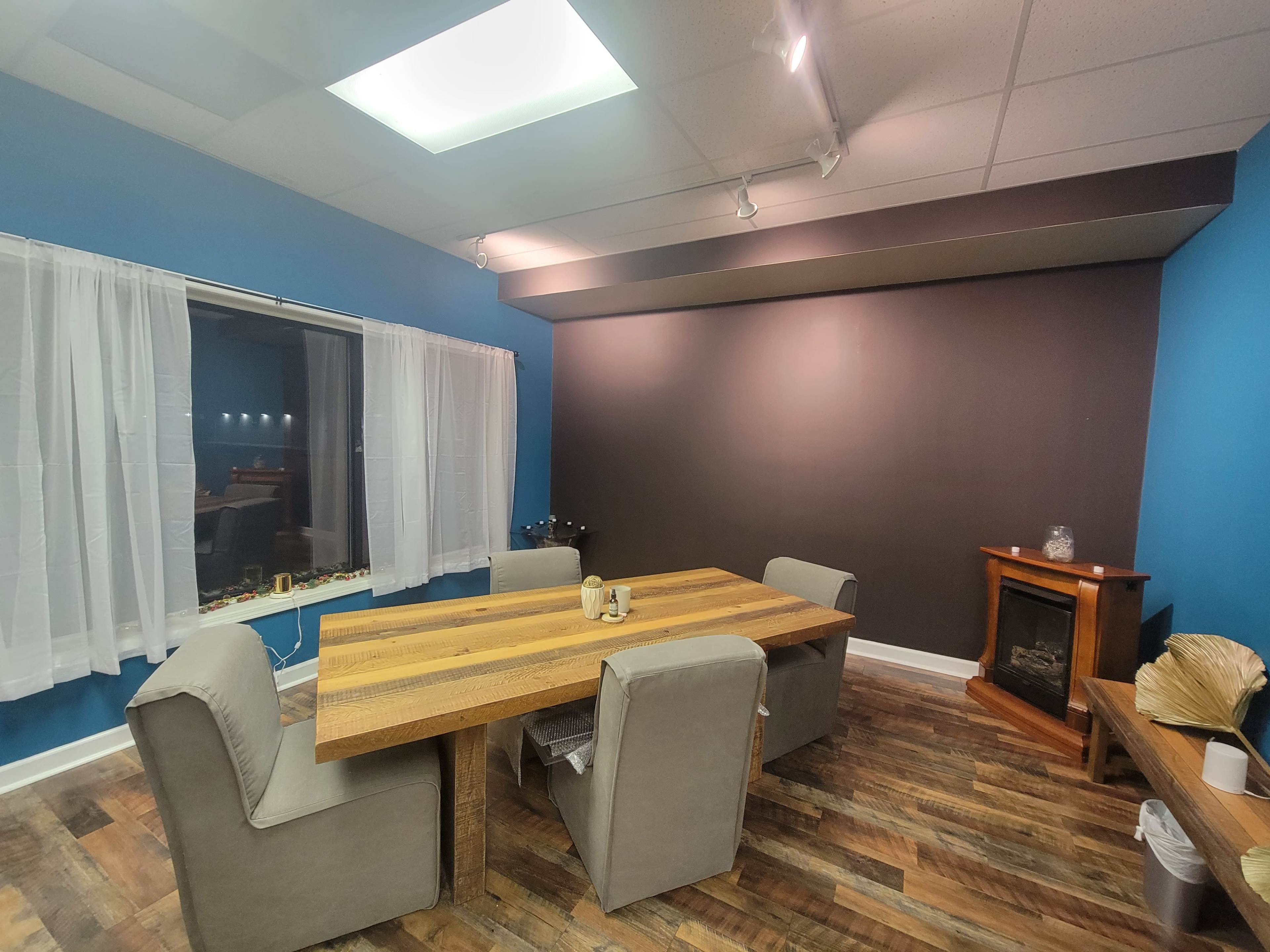 The image shows a modern room with a wooden dining table surrounded by upholstered chairs, a cozy fireplace in the corner, and a large window with sheer curtains.