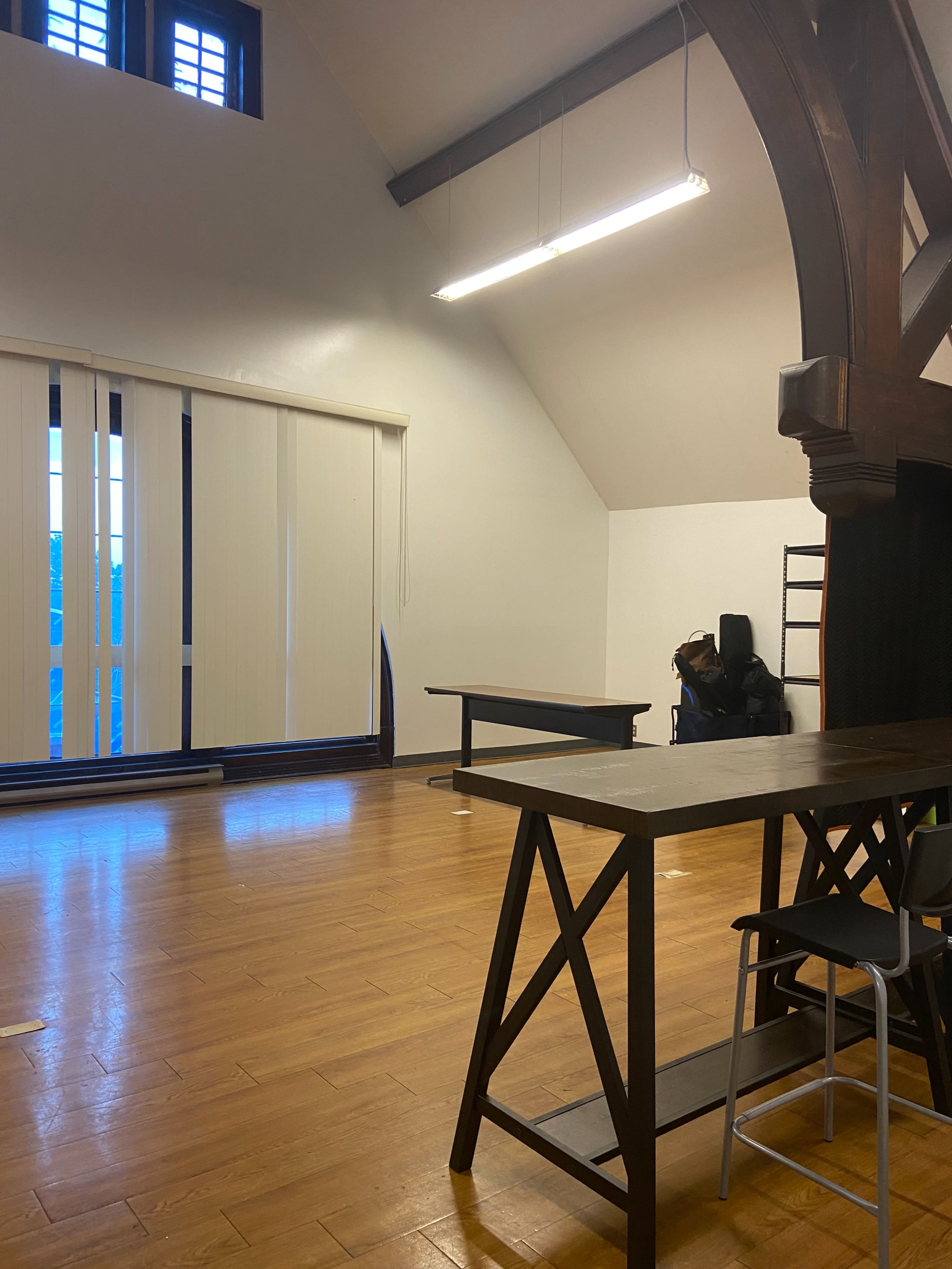 A spacious room with wooden floors, featuring a large window covered by vertical blinds, two tables, and a ladder against the wall.
