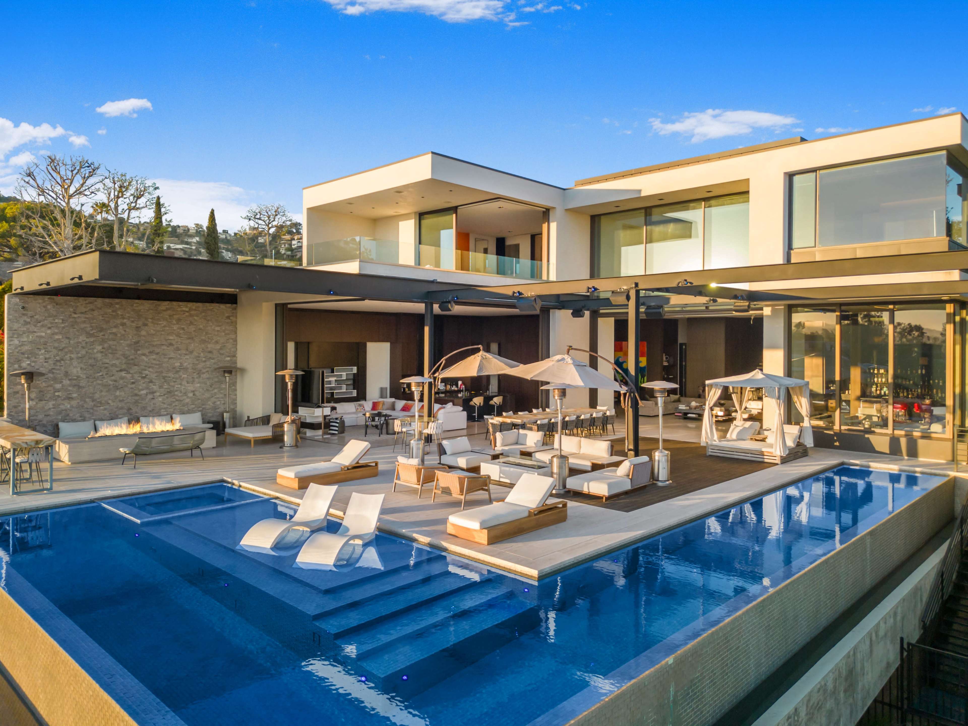 The image shows a modern luxury home with a large swimming pool and lounge area, situated in a scenic outdoor setting.