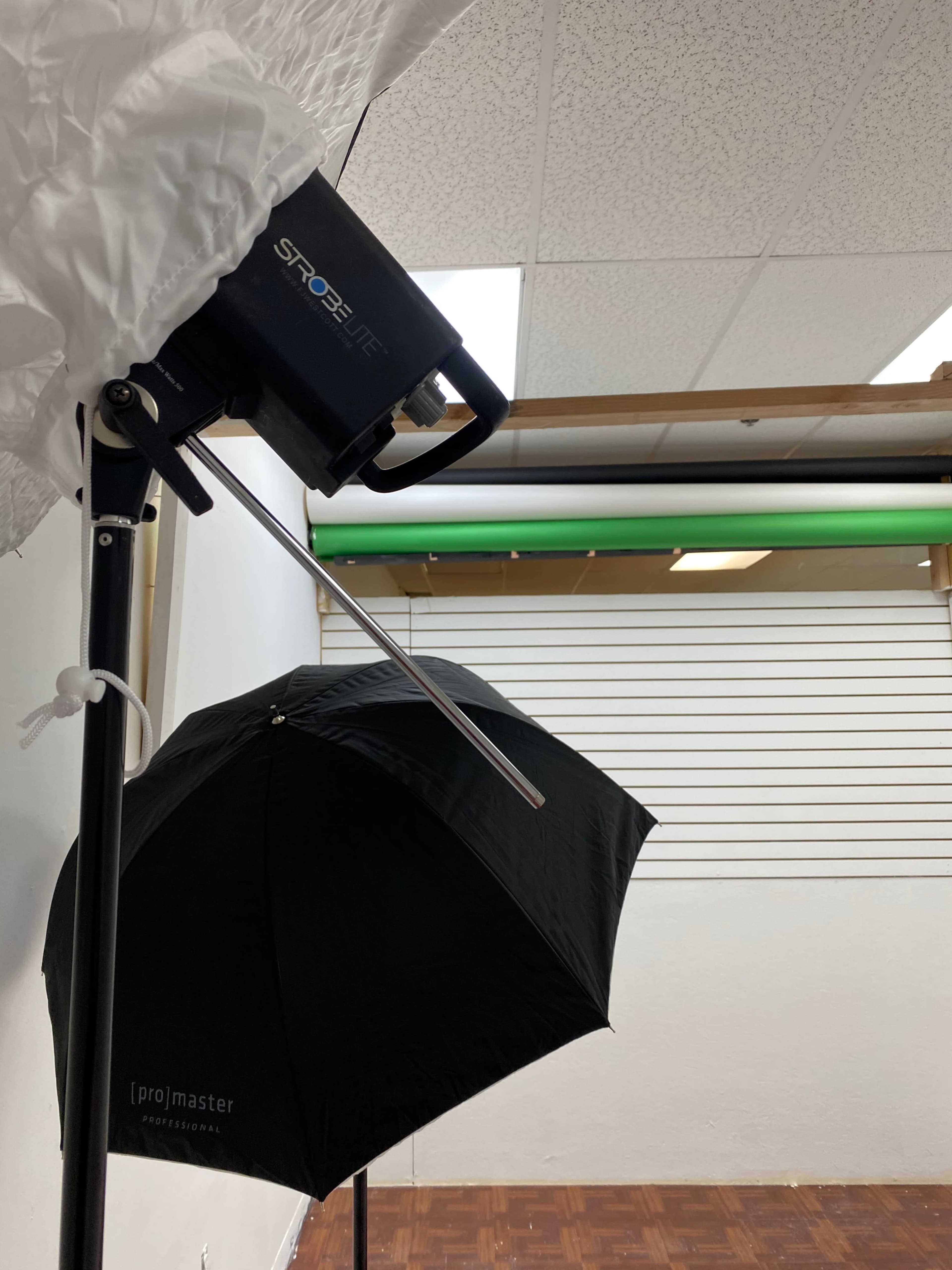A photography setup features a strobe light mounted on a stand near a black umbrella, with a backdrop displaying green and white horizontal lines.