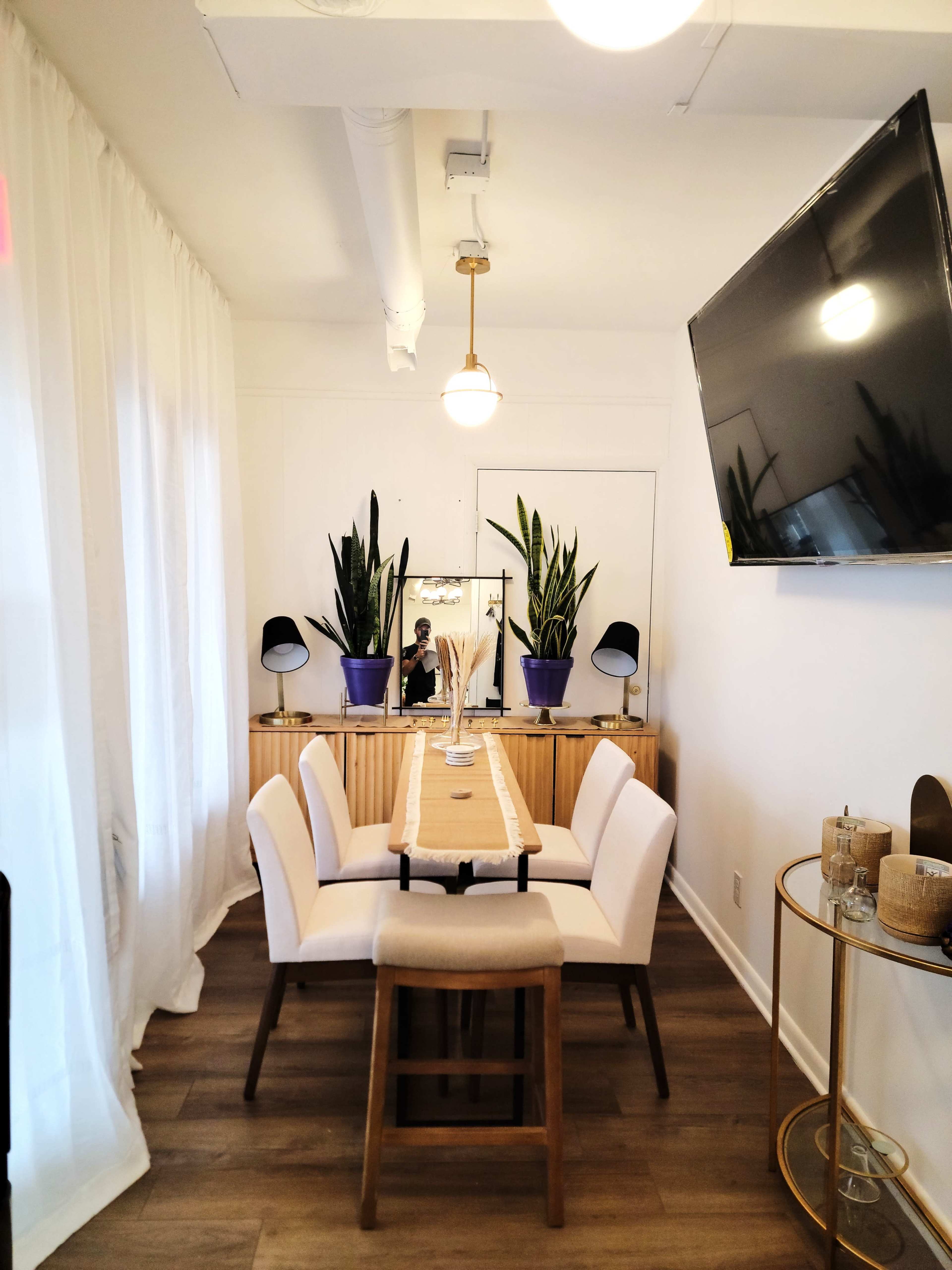 A dining area features a wooden table surrounded by six upholstered chairs, with a mirror and potted plants on a sideboard, and a large television mounted on the wall.