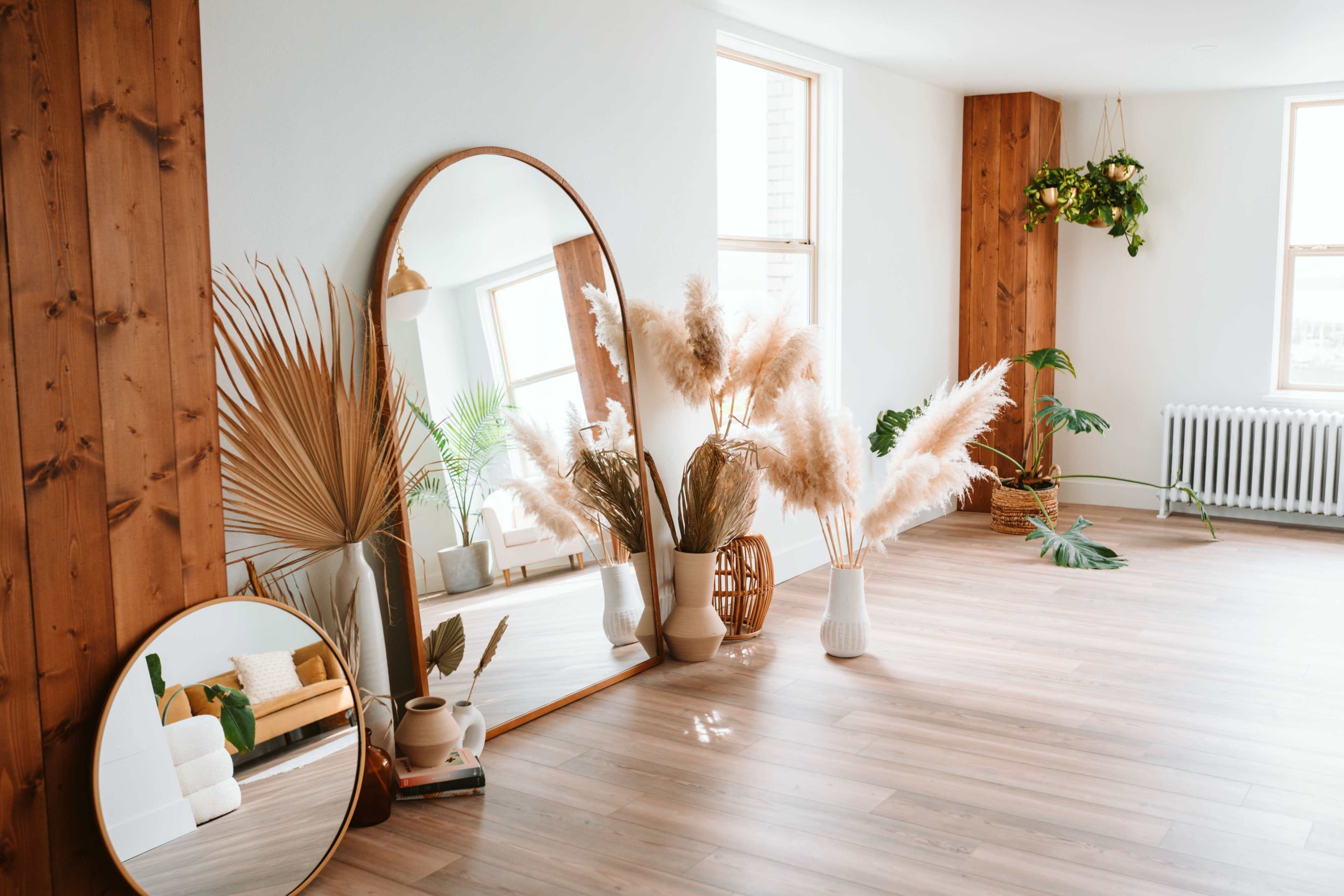 A bright room features large mirrors, decorative plants, and pampas grass arranged near wooden walls and large windows.