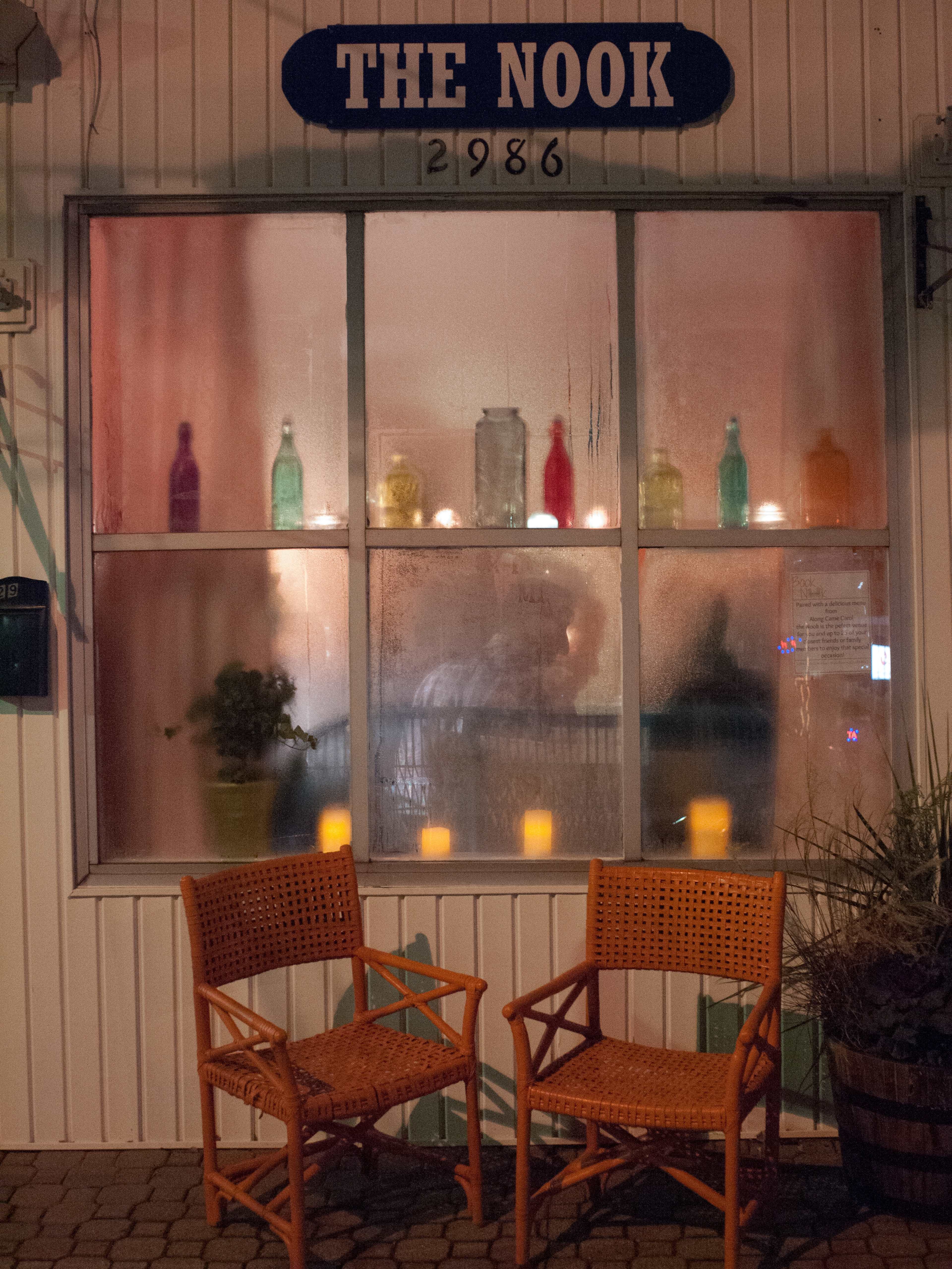 The image shows the exterior of a building named "The Nook," featuring a foggy window with colorful bottles displayed on the ledge and two wicker chairs positioned outside.