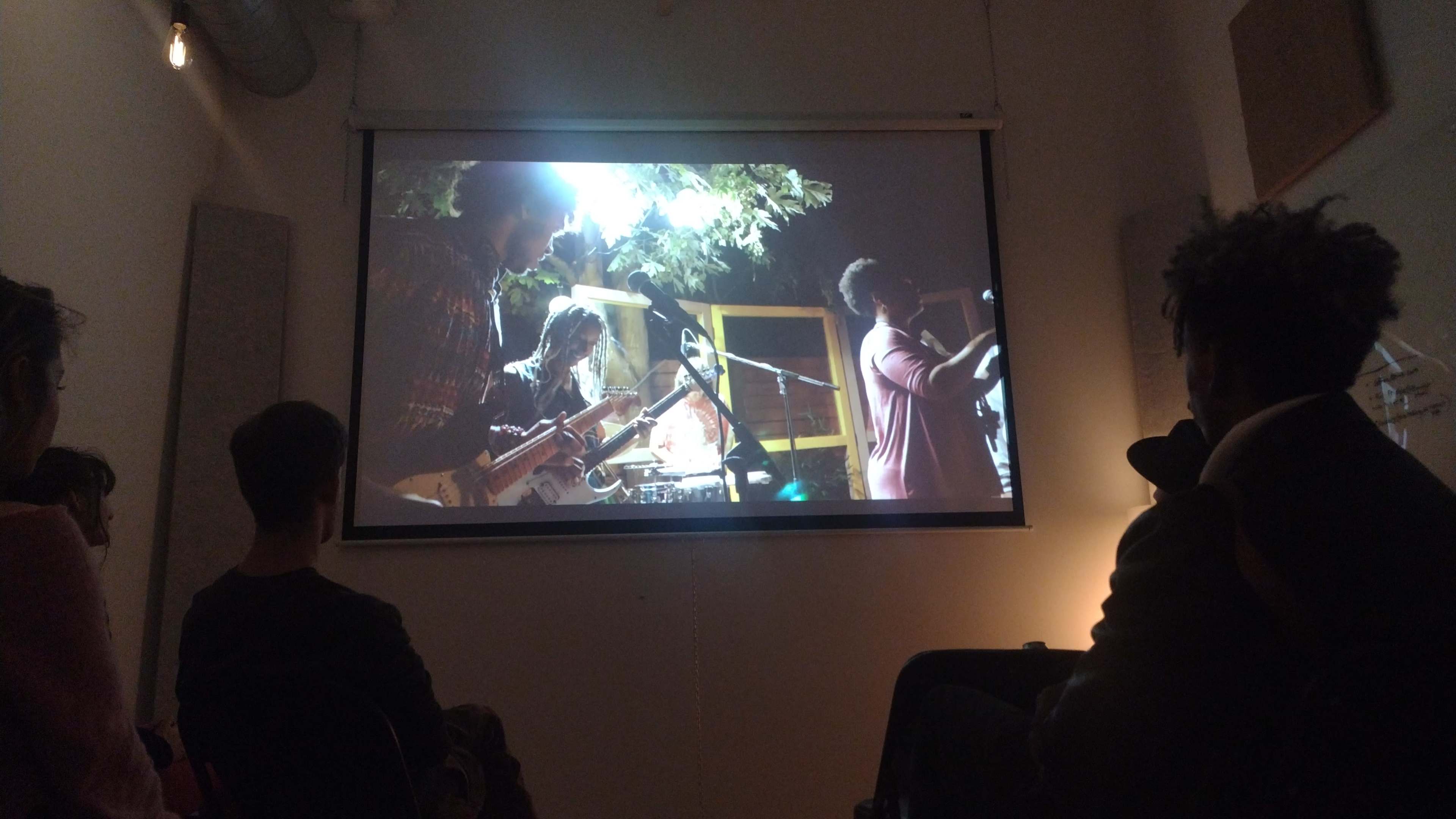A group of musicians performs on stage while an audience sits in front of a large screen displaying the performance.