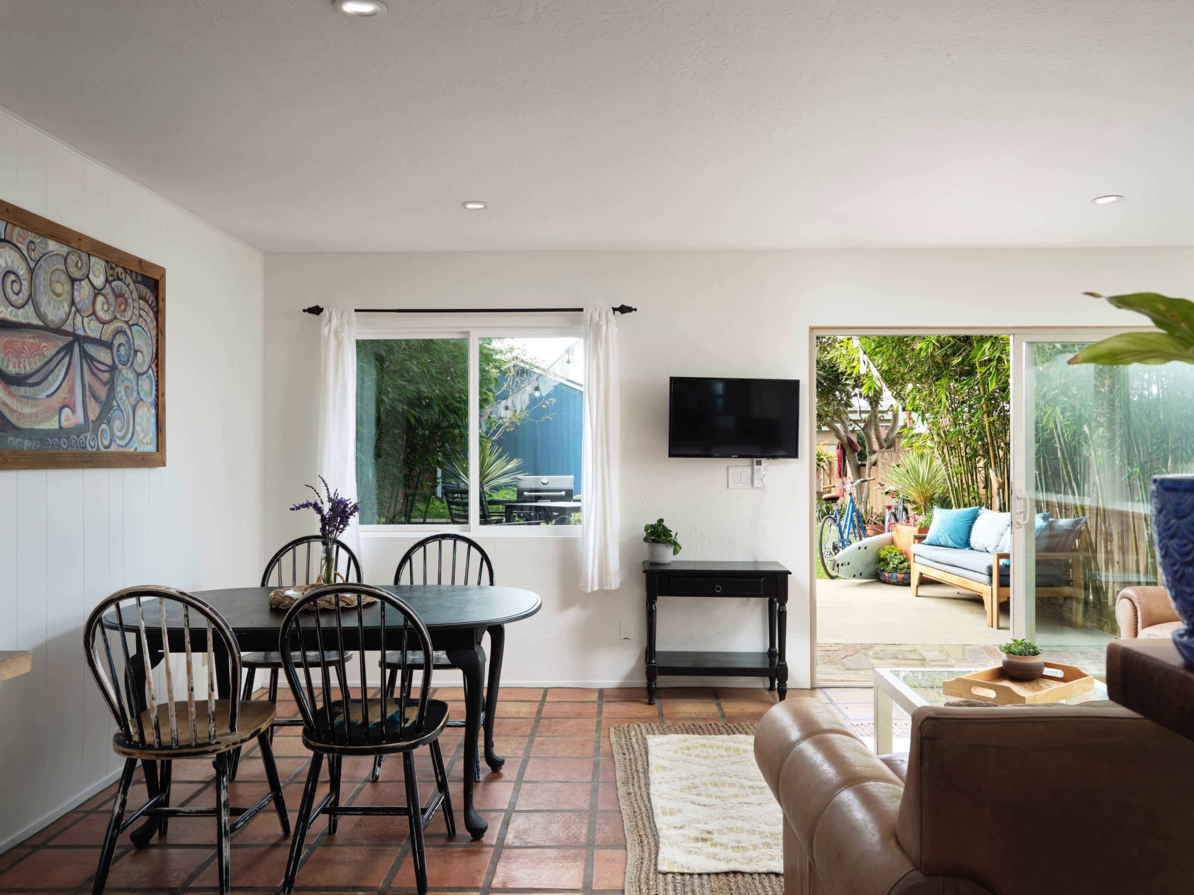 A cozy living space with a dining table, a television mounted on the wall, and large windows overlooking a garden area.