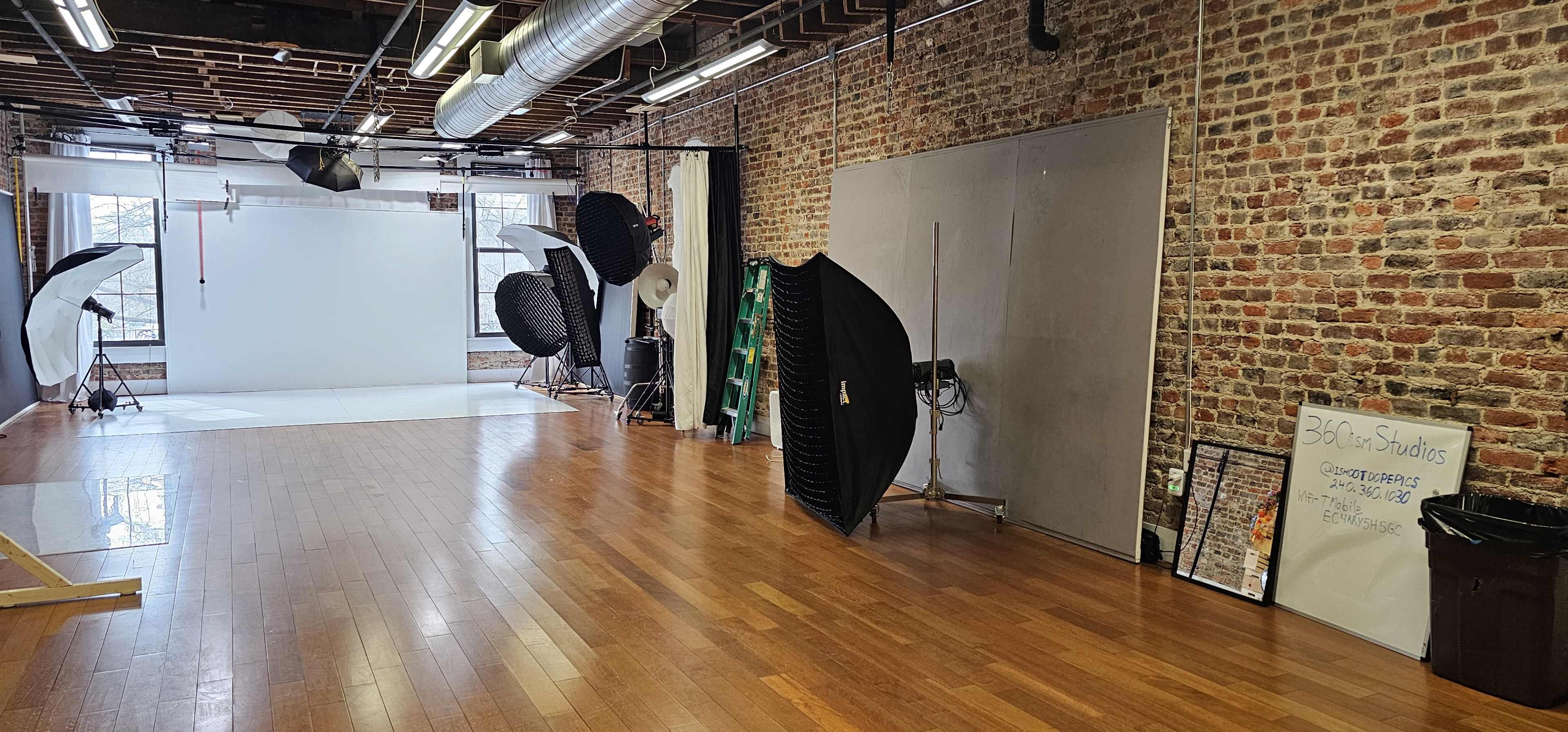 The image shows a spacious photography studio with a wooden floor, exposed brick walls, and various lighting equipment arranged along one side.