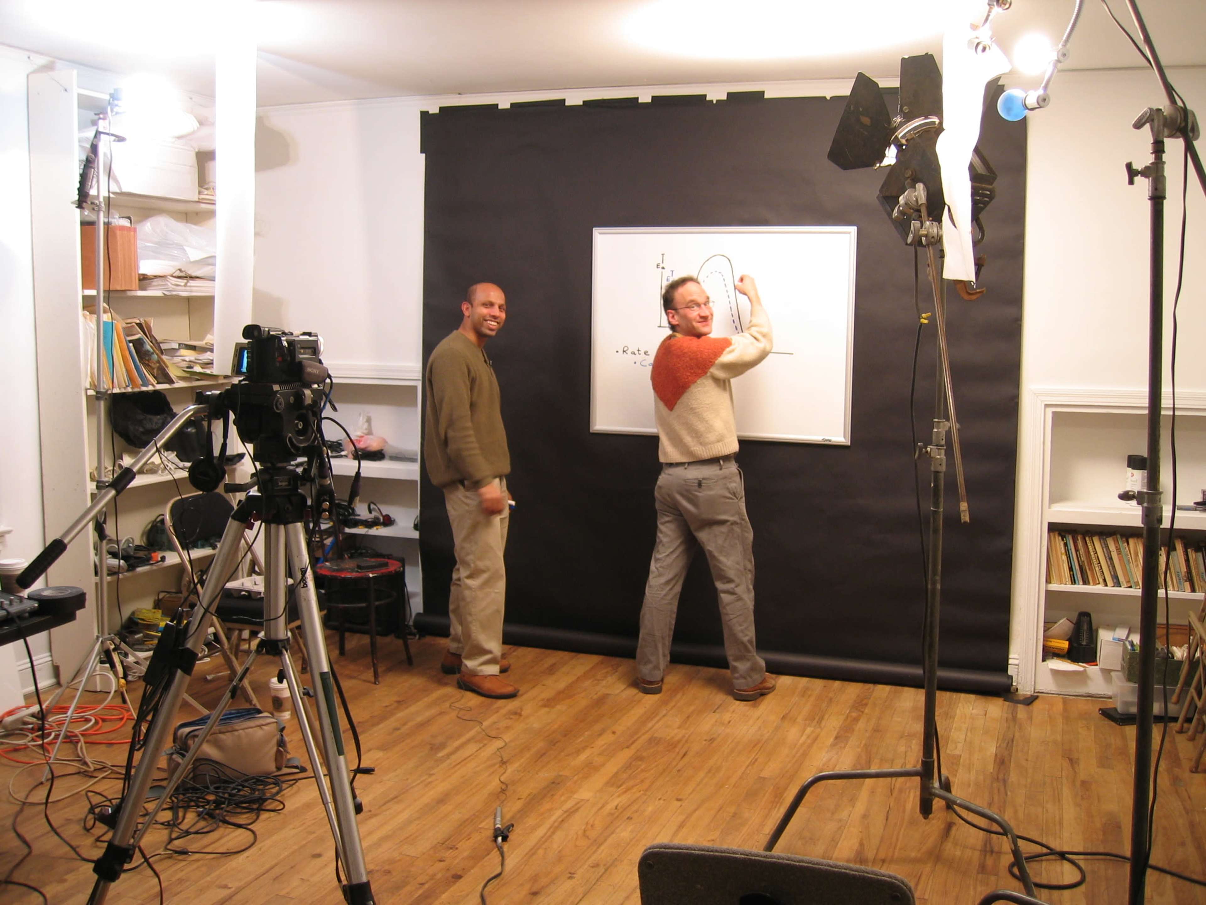 Two men stand in a studio in front of a black backdrop, one drawing on a whiteboard while the other looks on, with cameras and lights set up around them.