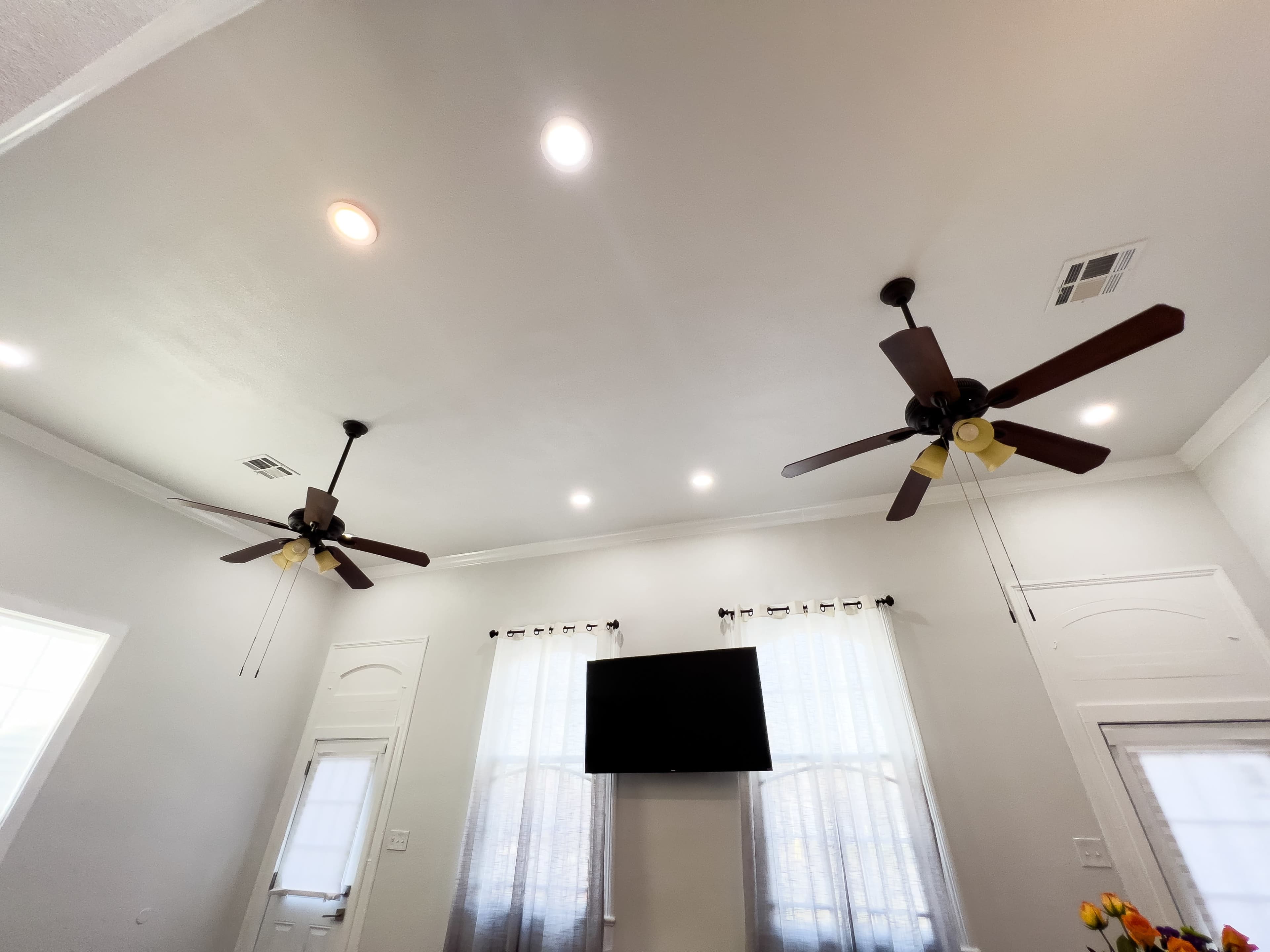 A room with a high ceiling featuring two ceiling fans, recessed lighting, and a wall-mounted television above a door.