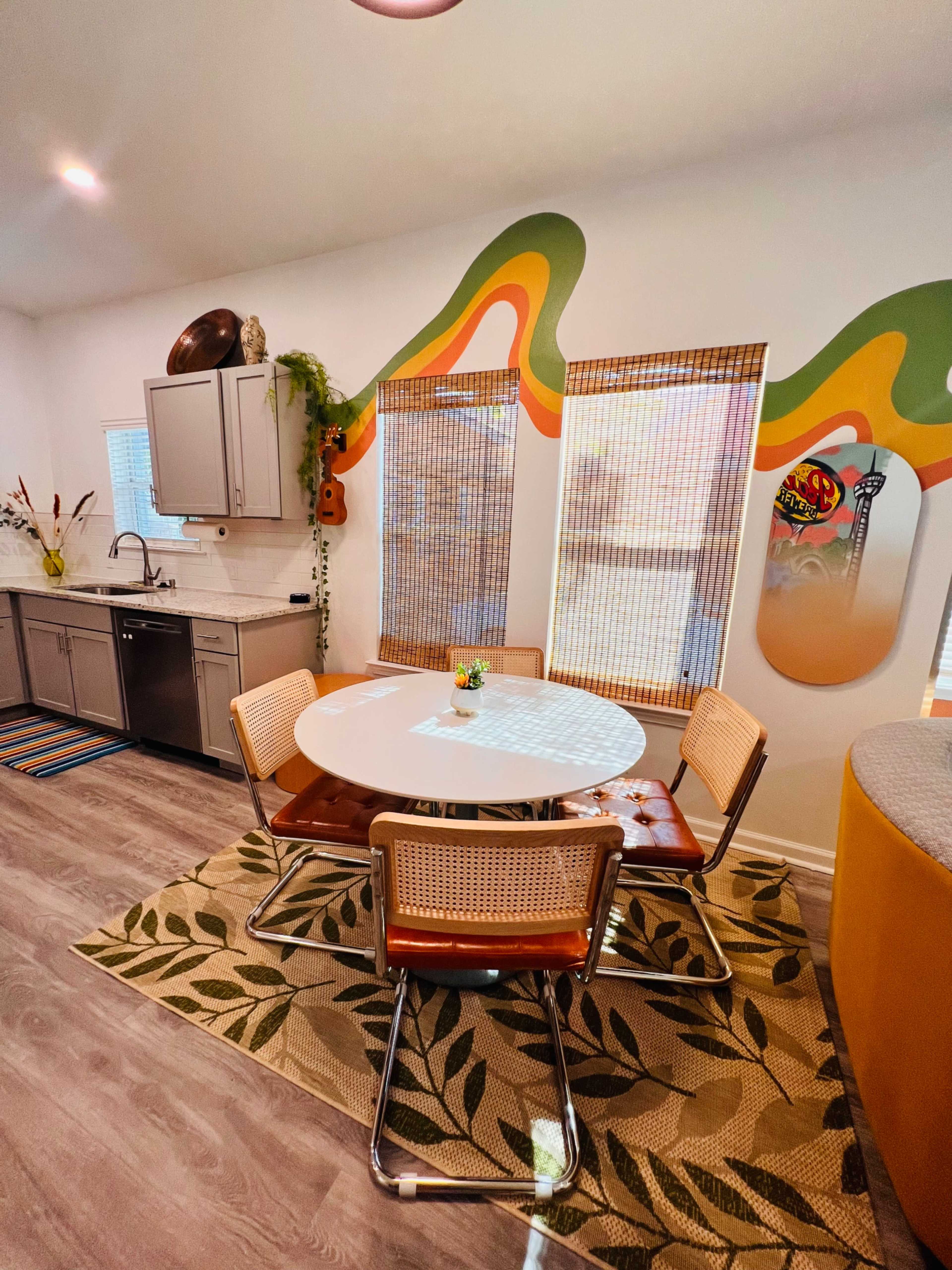 A modern kitchen features a round white dining table surrounded by four chairs on a patterned rug, with colorful wall decor and large windows covered by bamboo shades.