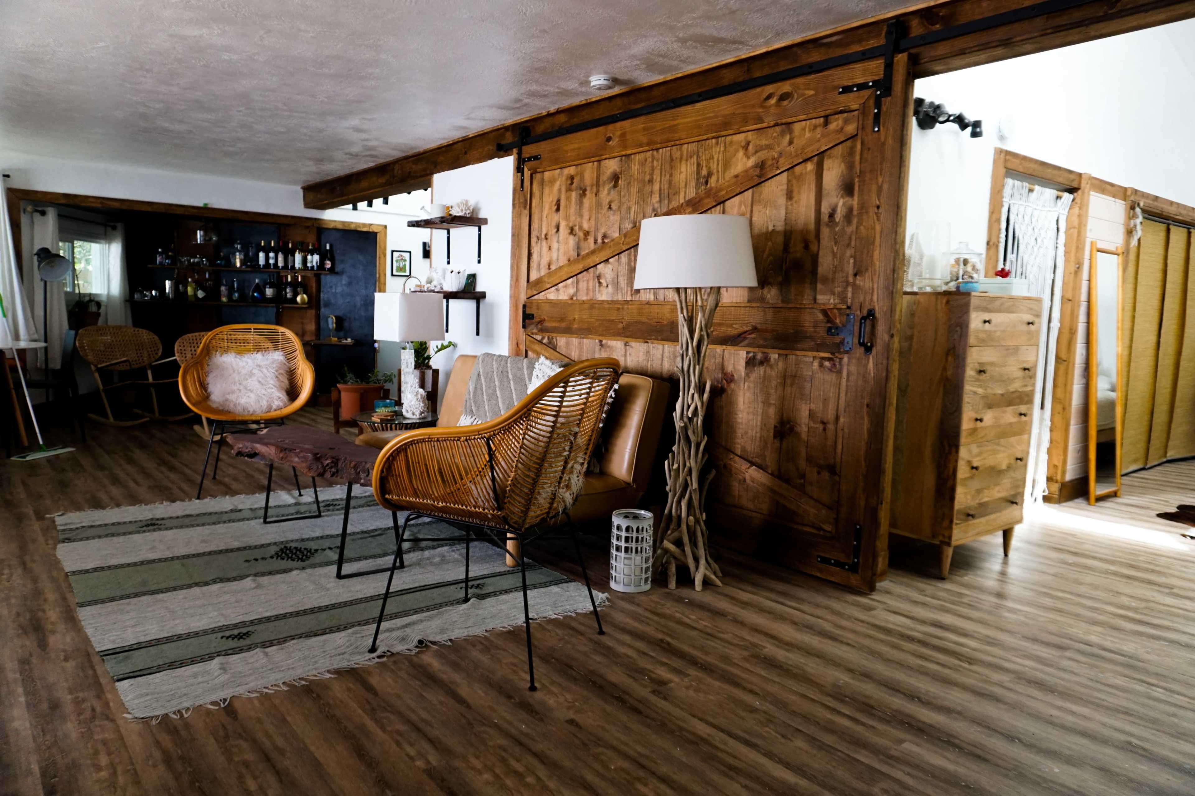 The image shows a modern living room with wooden accents, featuring a pair of woven chairs, a decorative table, and a large sliding barn door.