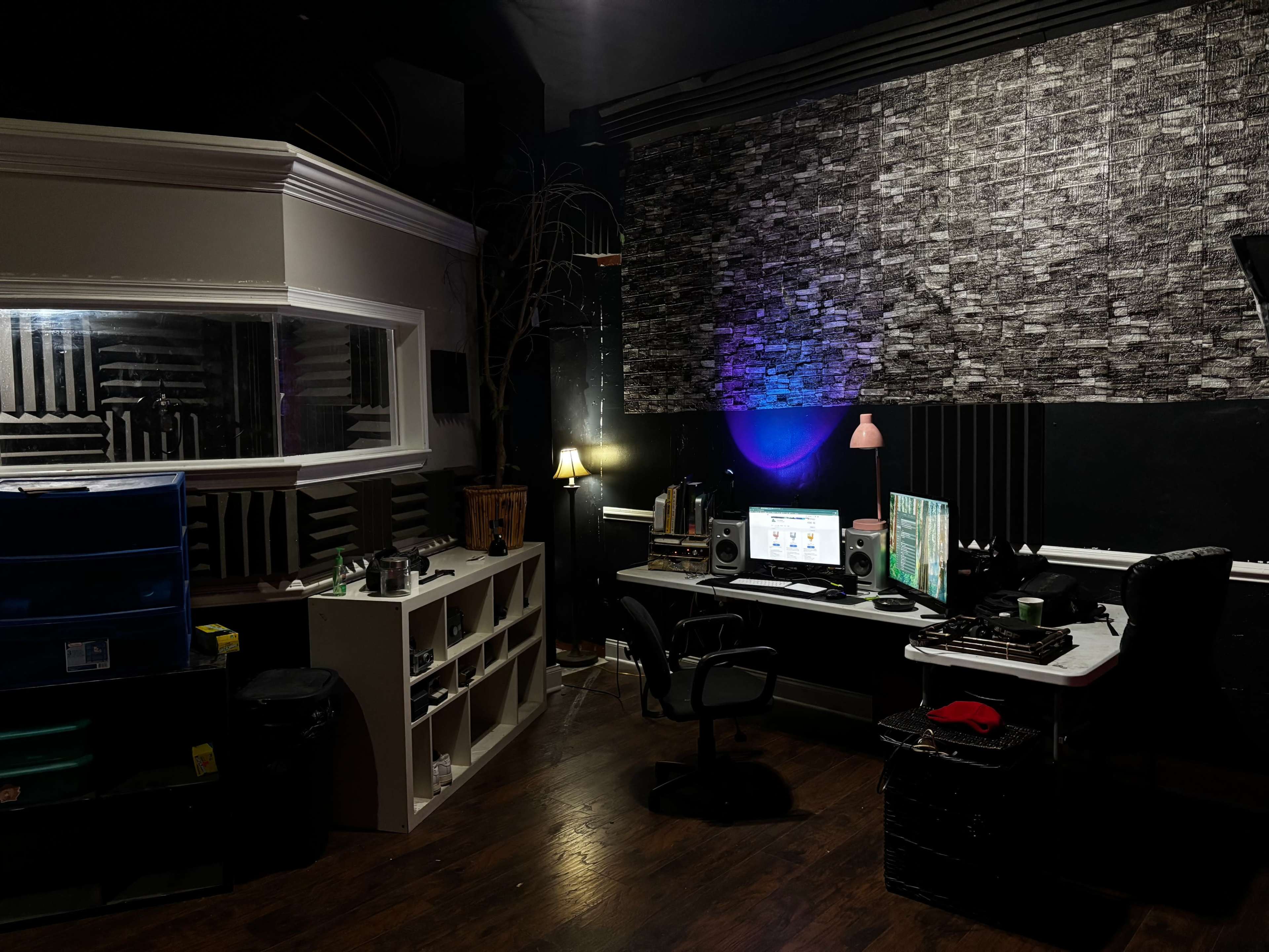 The image shows a dimly lit recording studio featuring a desk with audio equipment, a computer, and soundproofing materials on the walls.