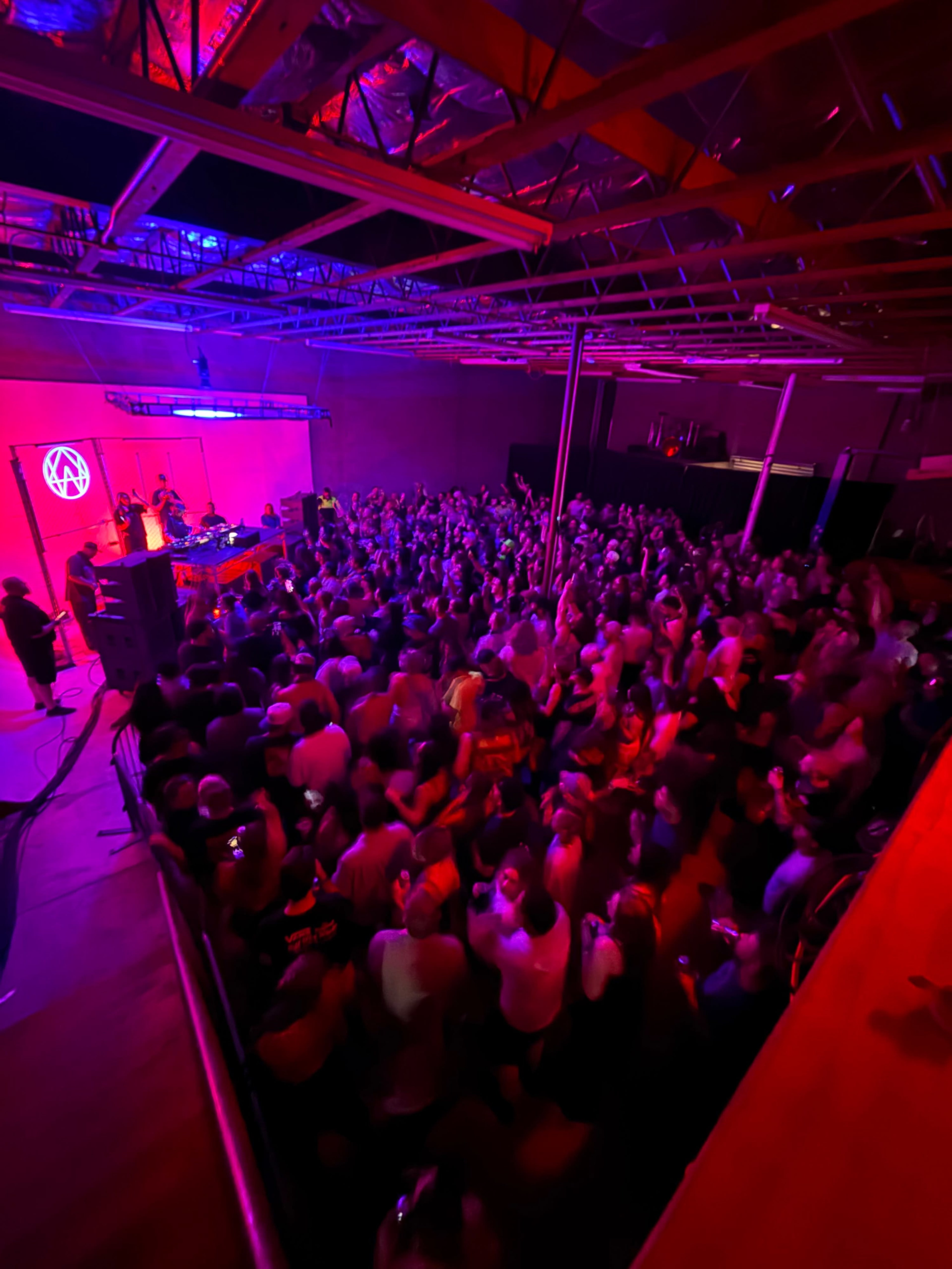 A large crowd enjoys a performance in a dimly lit venue with colorful lighting.