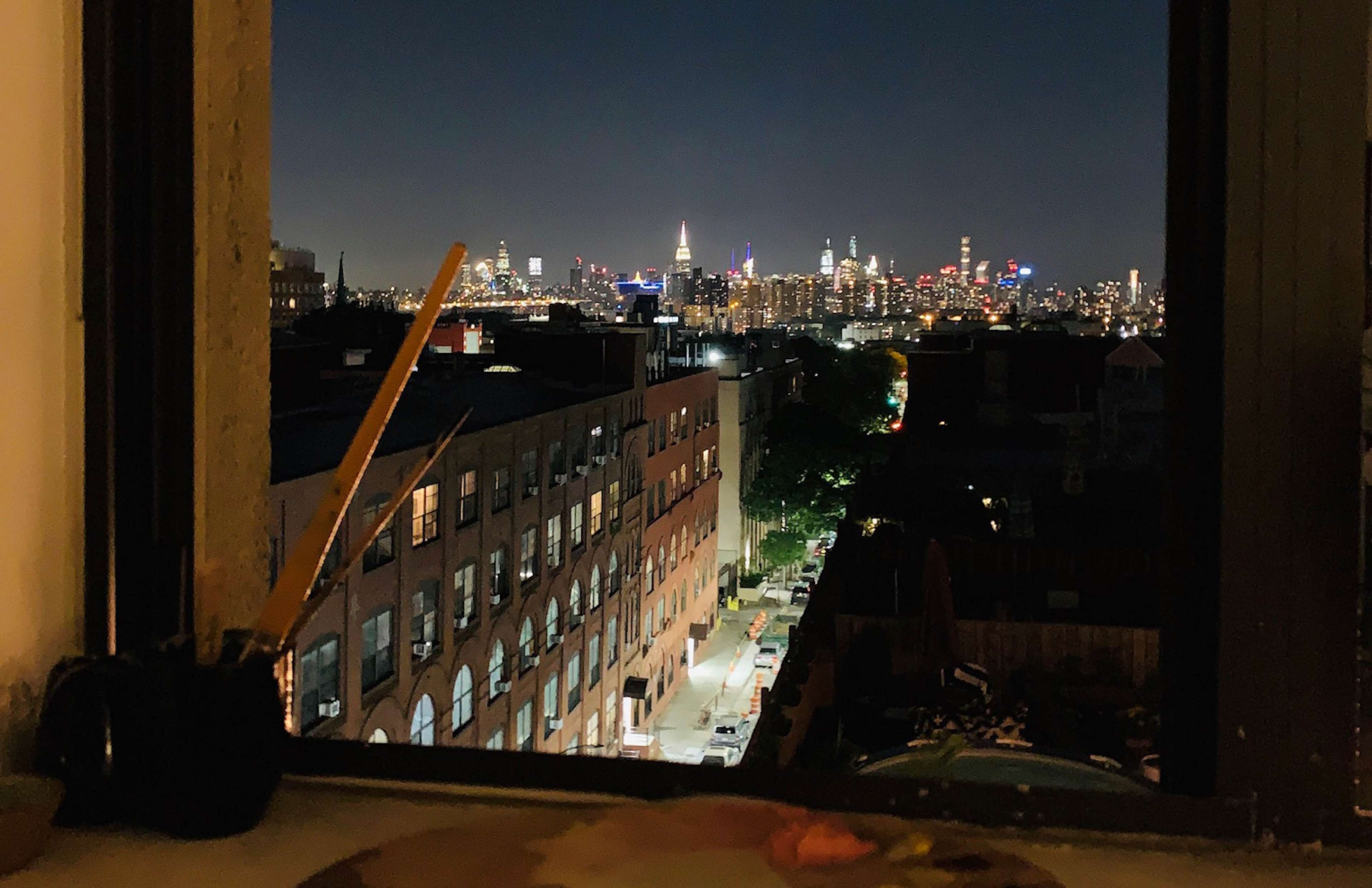 A nighttime view of a city skyline, featuring tall buildings and lights, seen from an open window with art supplies in the foreground.