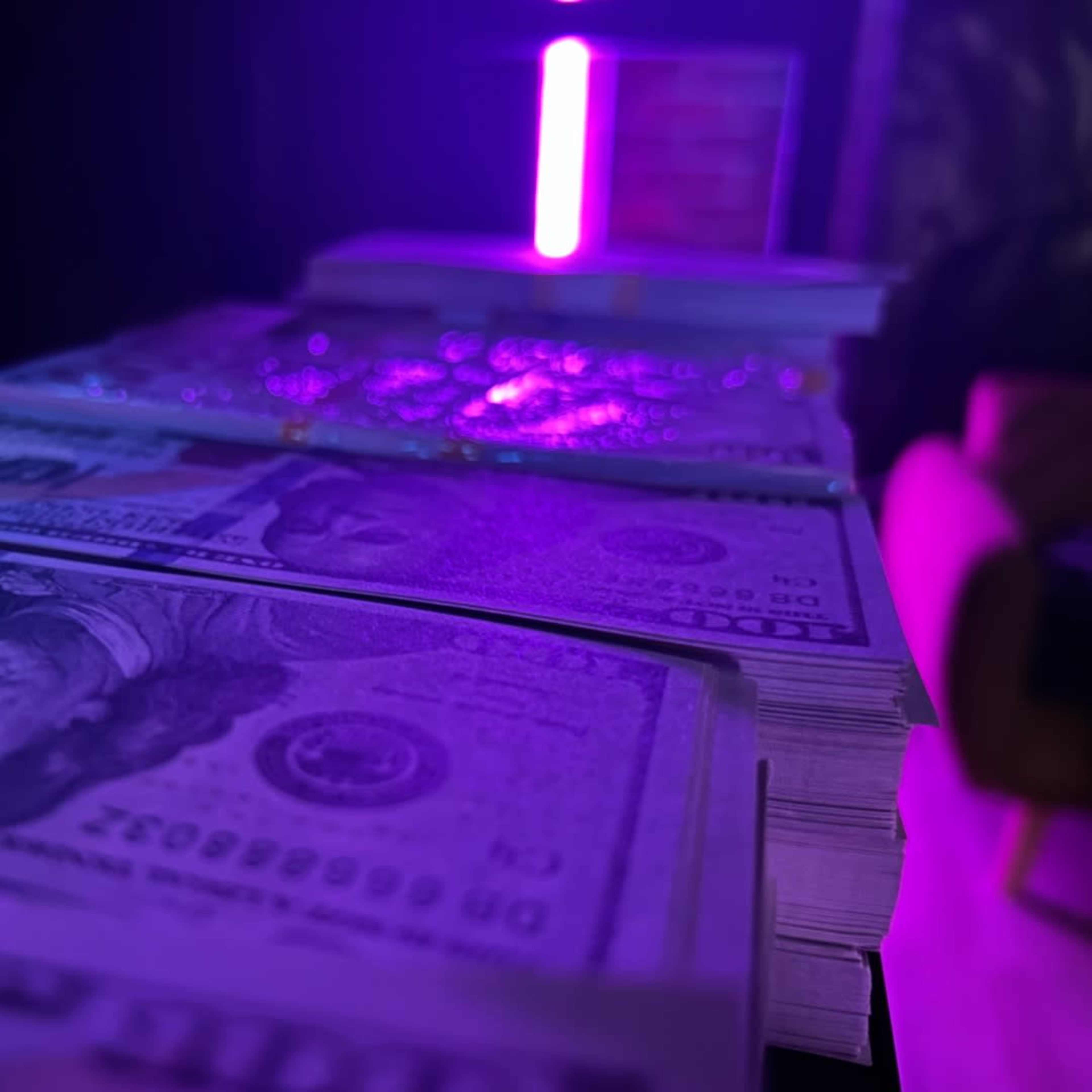 A close-up of stacks of U.S. hundred-dollar bills illuminated by a purple light.