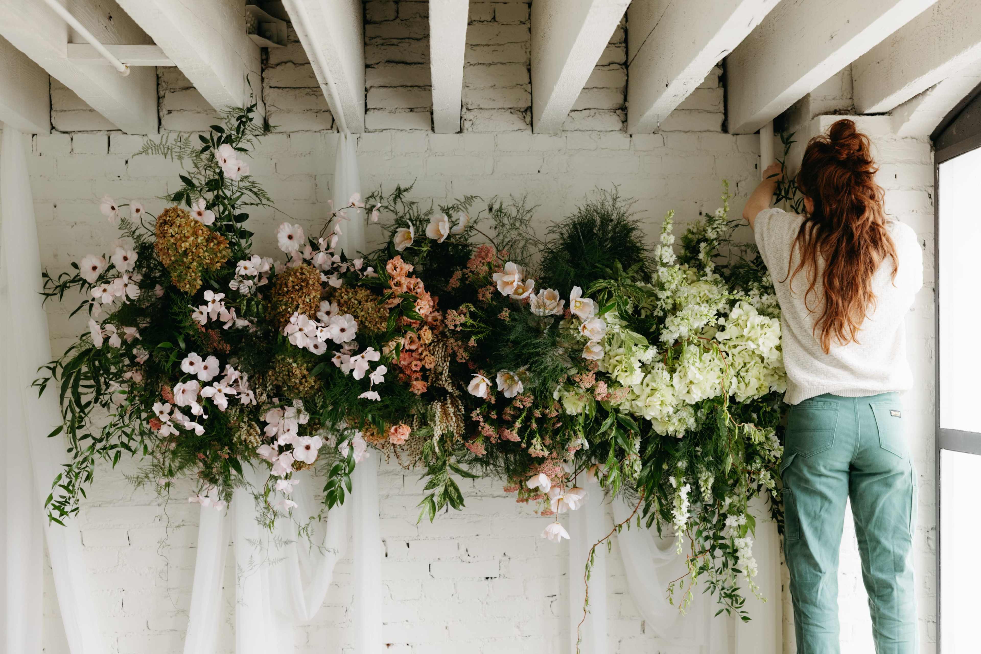 A person arranges a large floral display on a white brick wall beneath exposed ceiling beams.