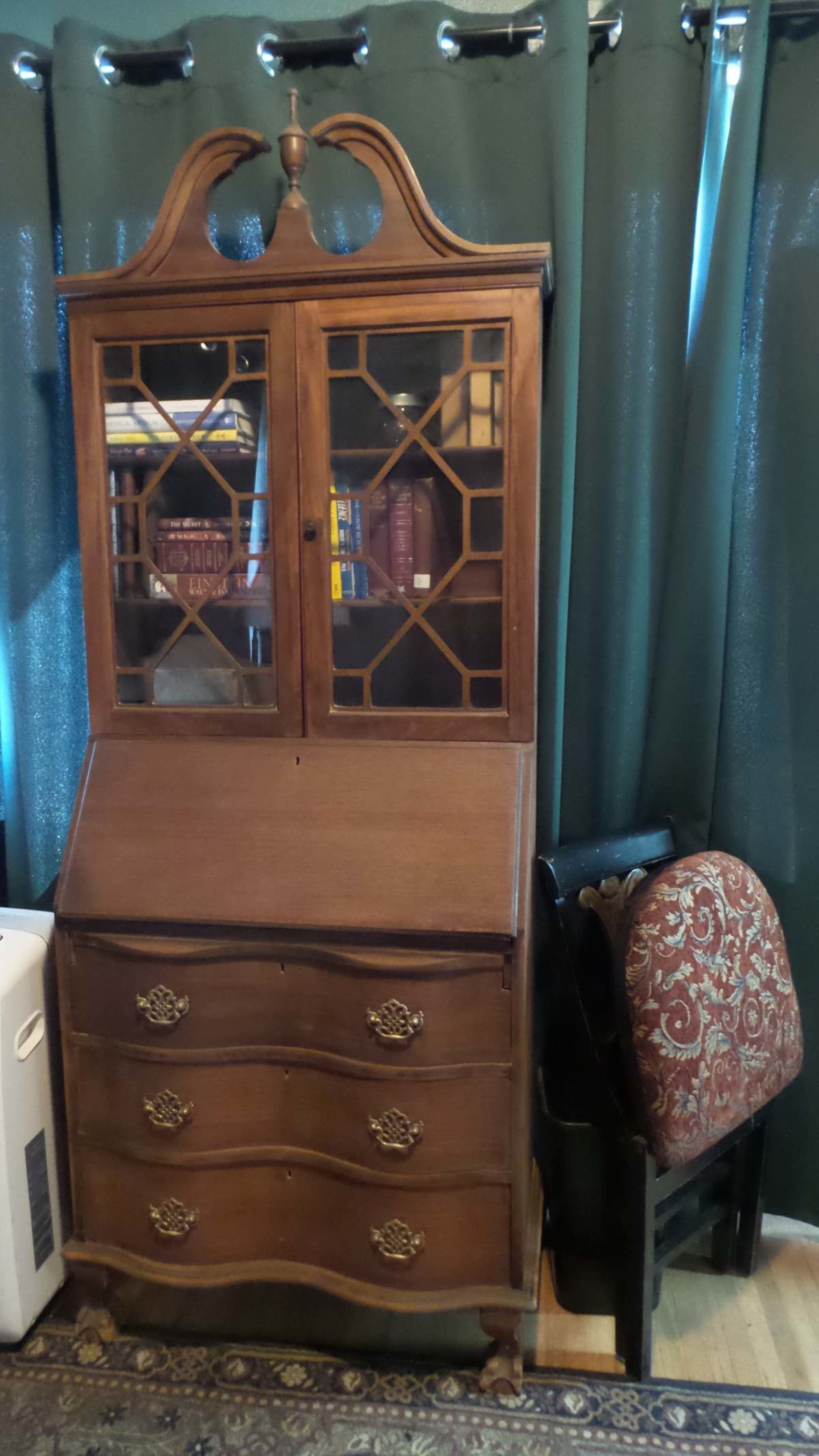 A wooden secretary desk with glass doors sits alongside a patterned upholstered chair on a hardwood floor, against a backdrop of green curtains.