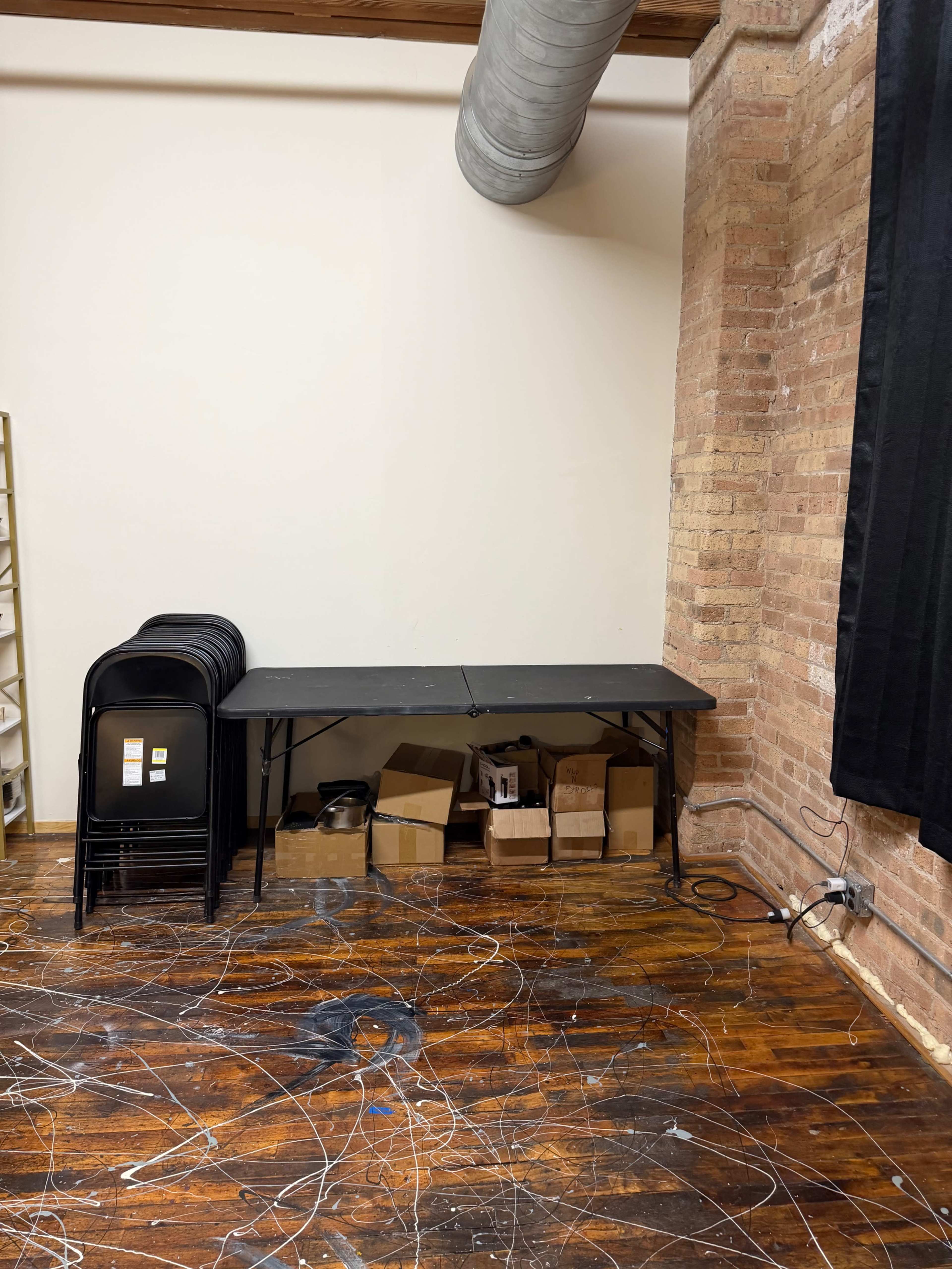 A black folding table is positioned against a brick wall, with stacked chairs behind it and cardboard boxes underneath.