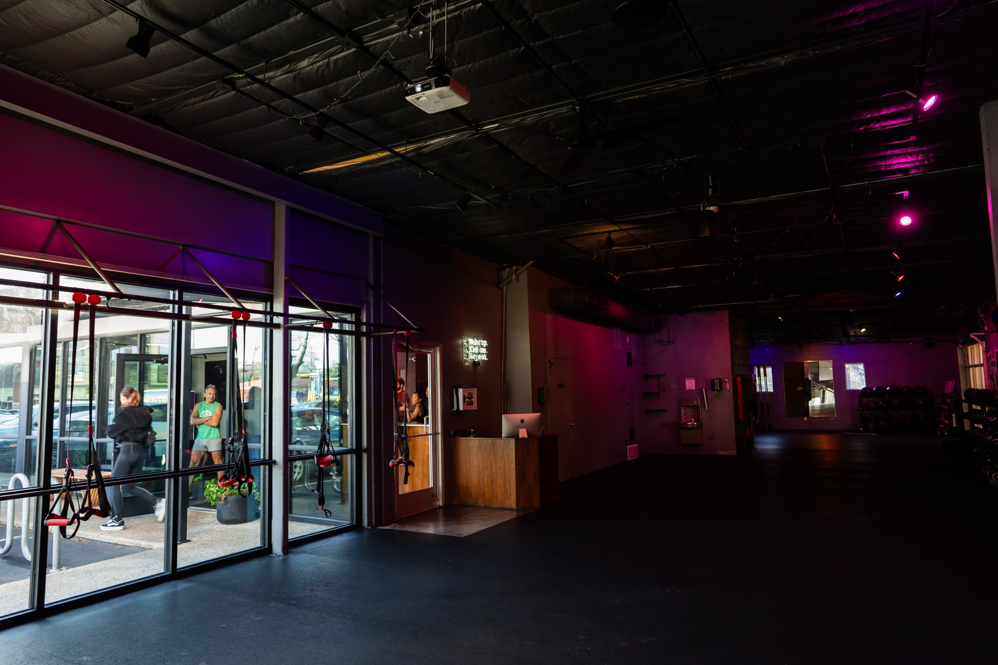 A spacious gym interior with a reception area, large glass doors, and a few people waiting outside.