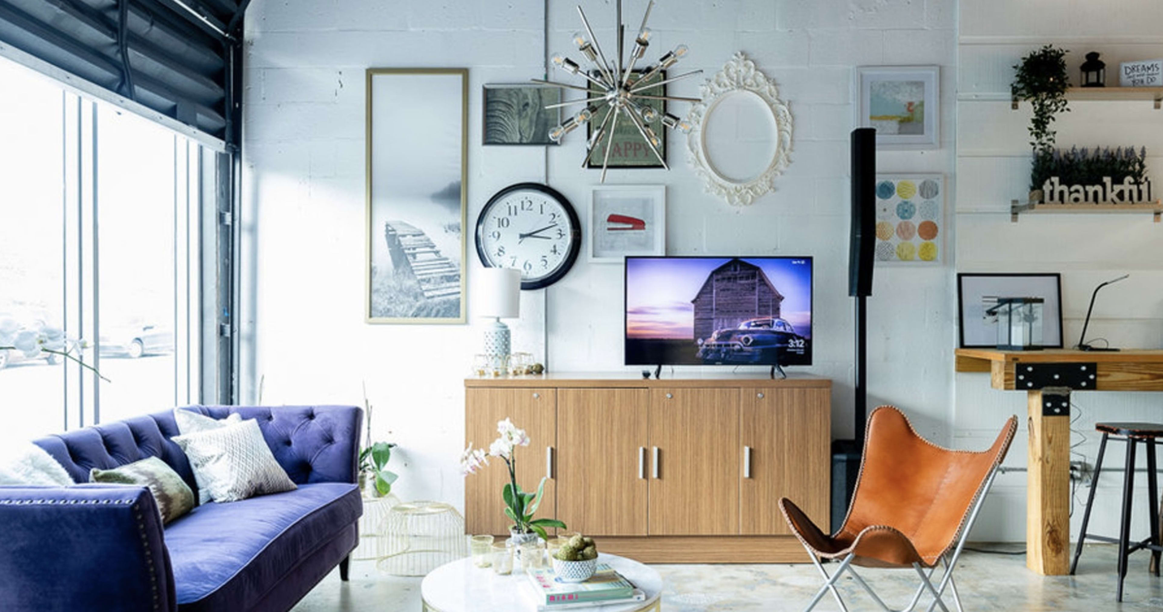 A modern living room featuring a purple sofa, a wooden media console with a television, an eclectic array of wall art, and a leather chair.