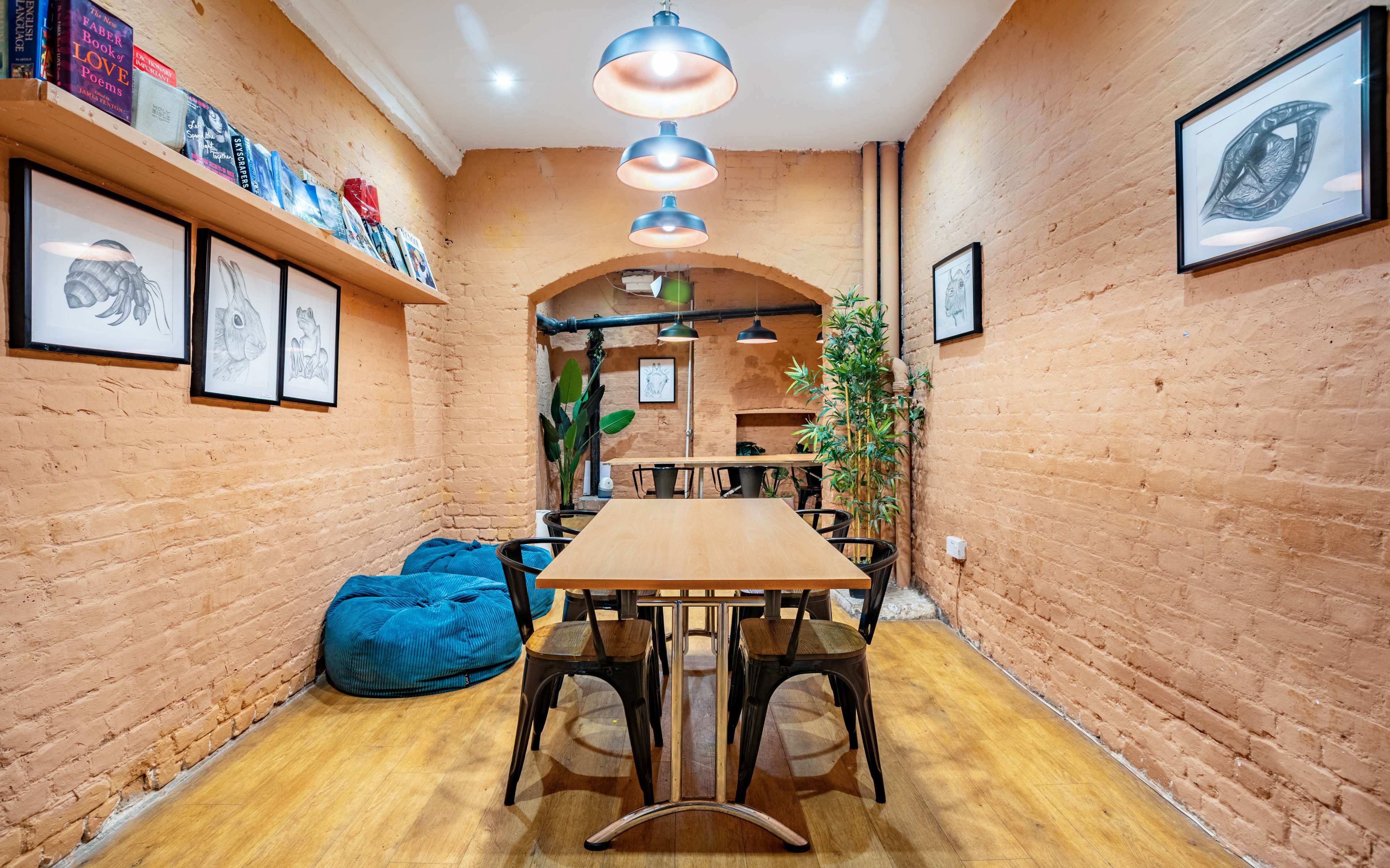 A cozy café interior features a long wooden table surrounded by black chairs, with books and framed artwork displayed on the walls.