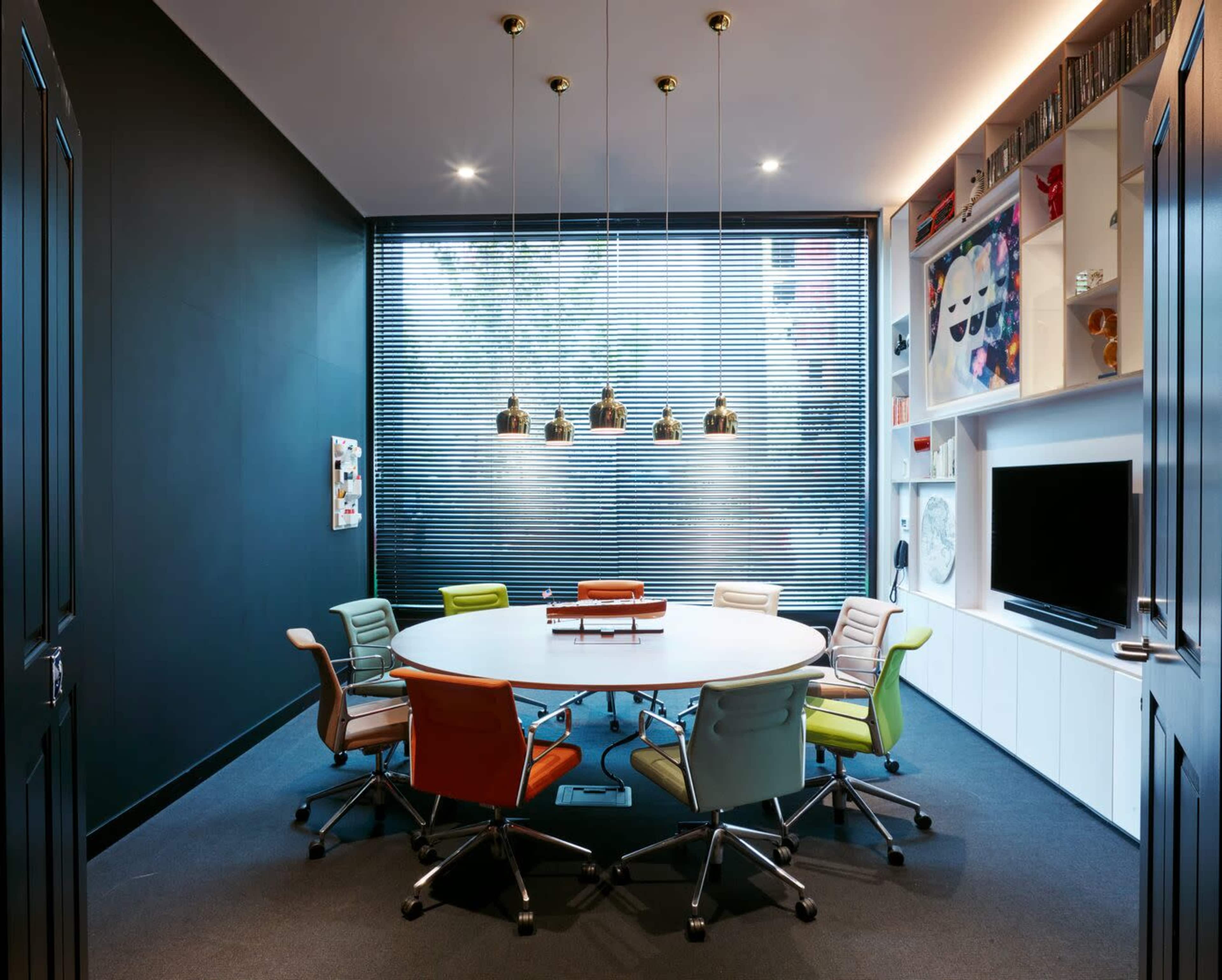 A modern conference room features a large round table surrounded by colorful chairs and a window with horizontal blinds.