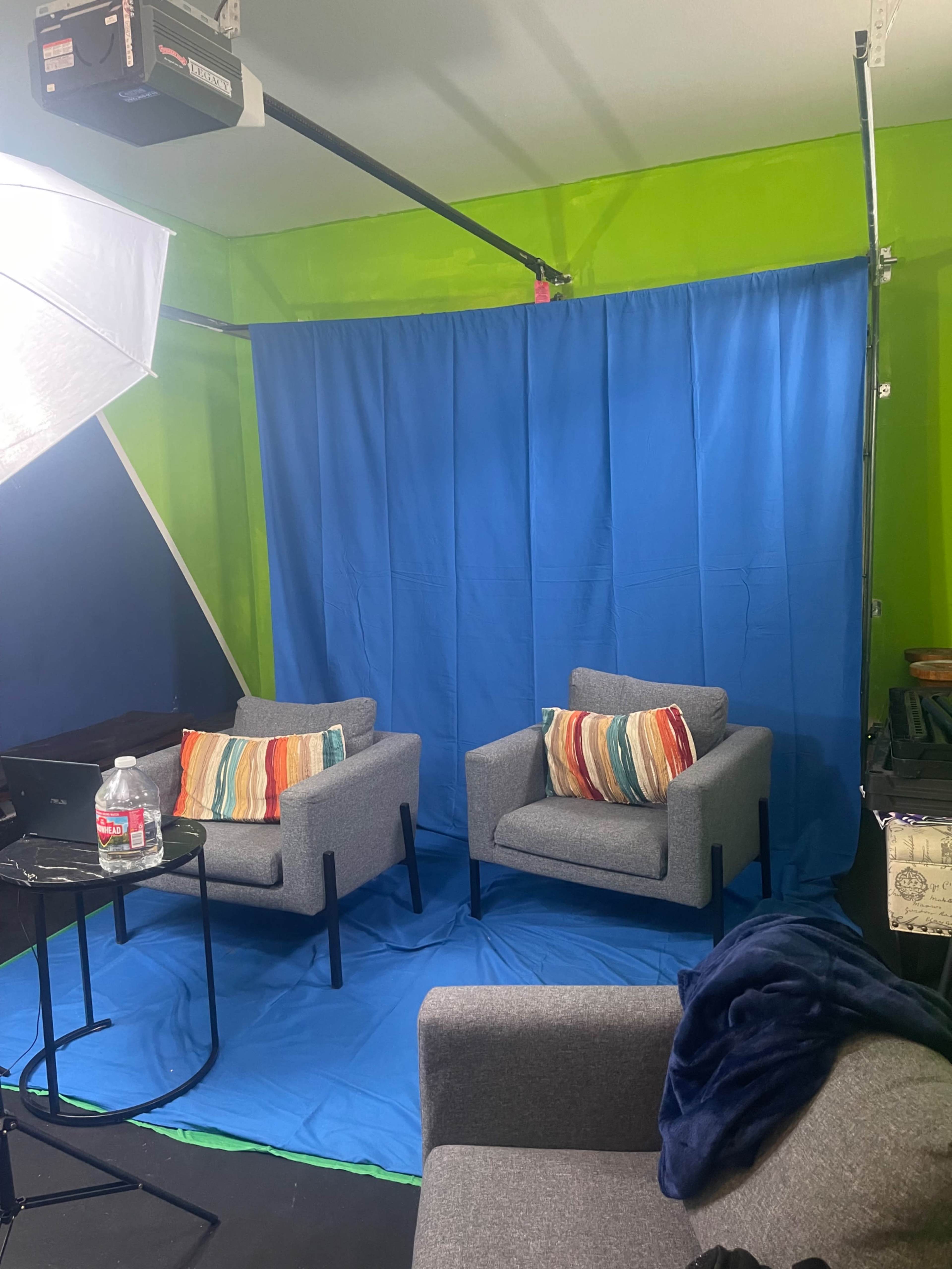 The image shows two chairs with decorative pillows in front of a blue curtain, set on a blue mat within a vividly painted green room.