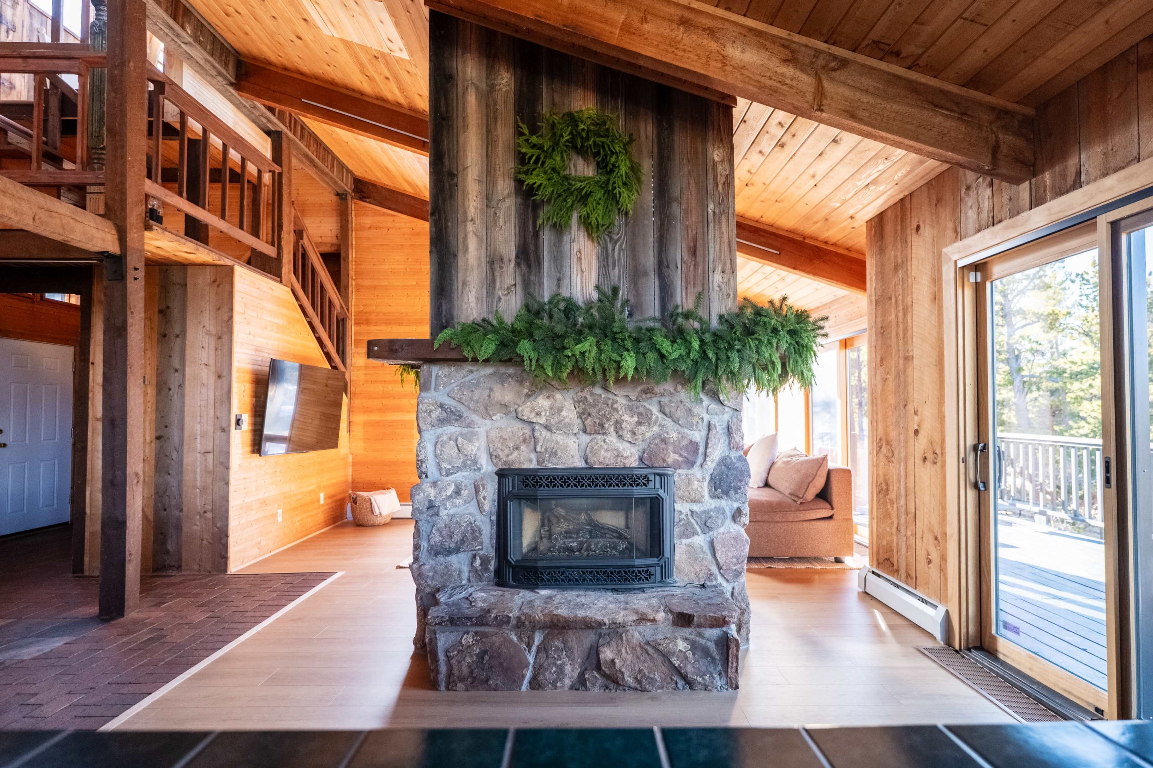 The image shows a rustic living room featuring a stone fireplace with a wooden mantel, surrounded by wooden walls and large windows that let in natural light.