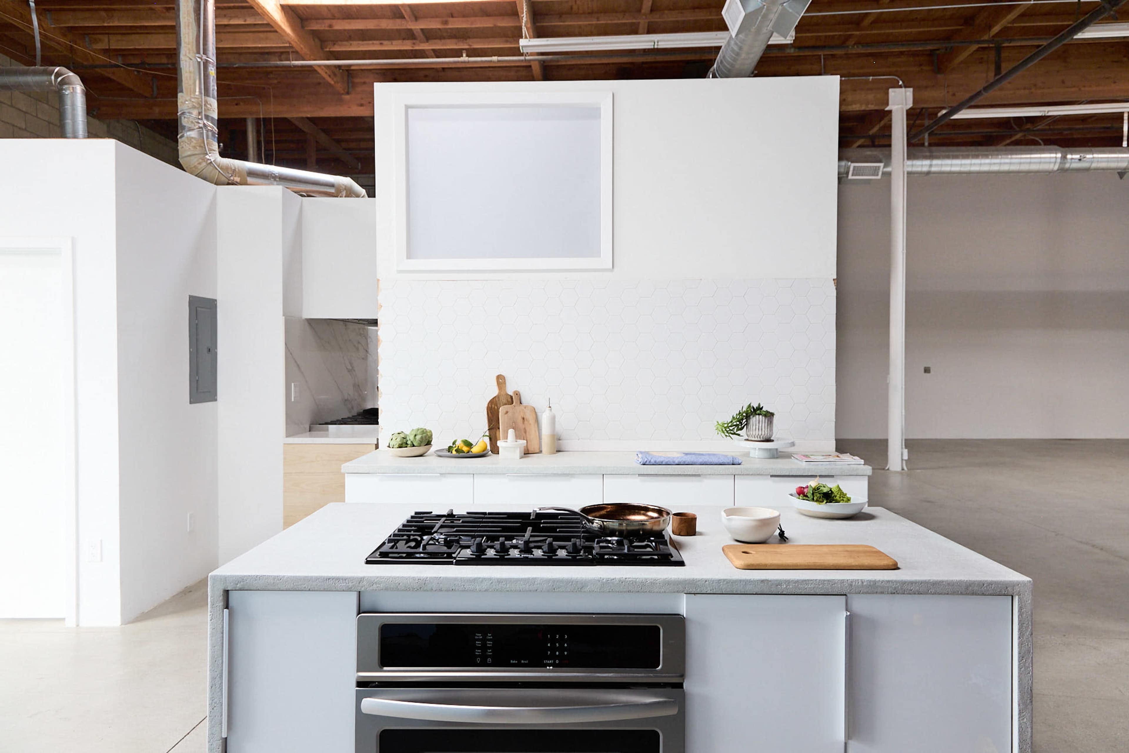A modern kitchen features a central island with a gas cooktop and minimalist decor, surrounded by white walls and large open space.